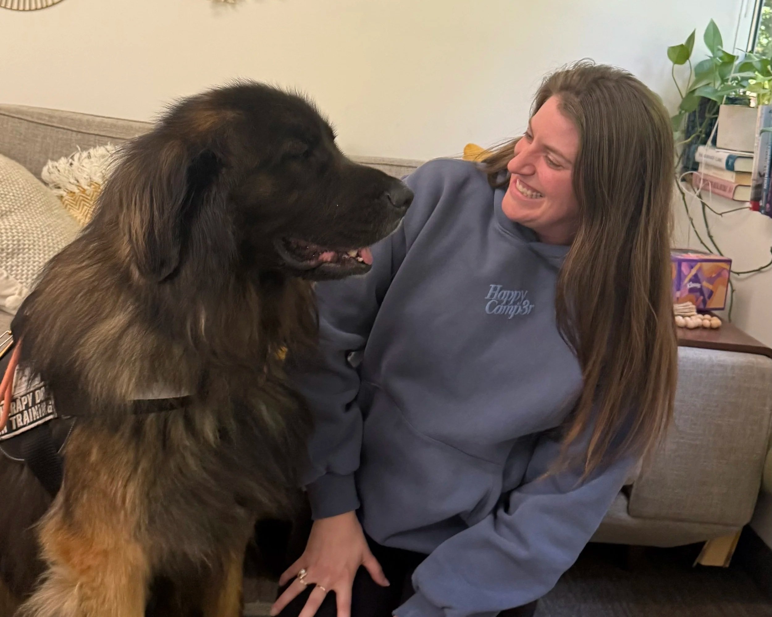 A woman with long brown hair smiling at a large black and brown dog that is standing close to her, indoors with a bookshelf and a potted plant in the background.