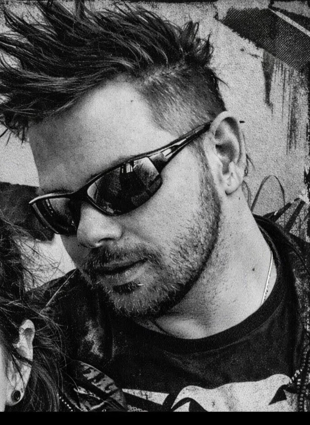 A close-up of JC Pass with spiked hair, sunglasses, and a beard, looking down, in black and white.