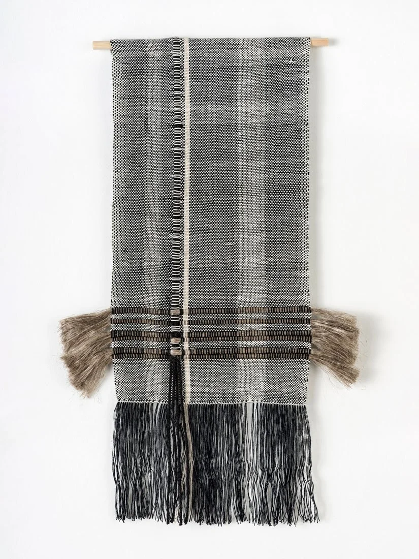 Black and white woven textile wall hanging with fringe at the bottom and linen tassel accents on the sides, mounted on a light-colored wooden dowel.