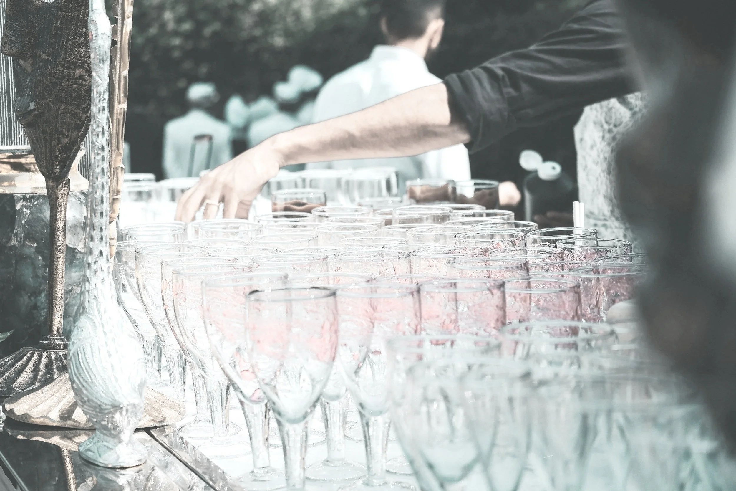 A person reaching out to glassware at an outdoor event or celebration, with several empty glasses and some decorated items on a table.