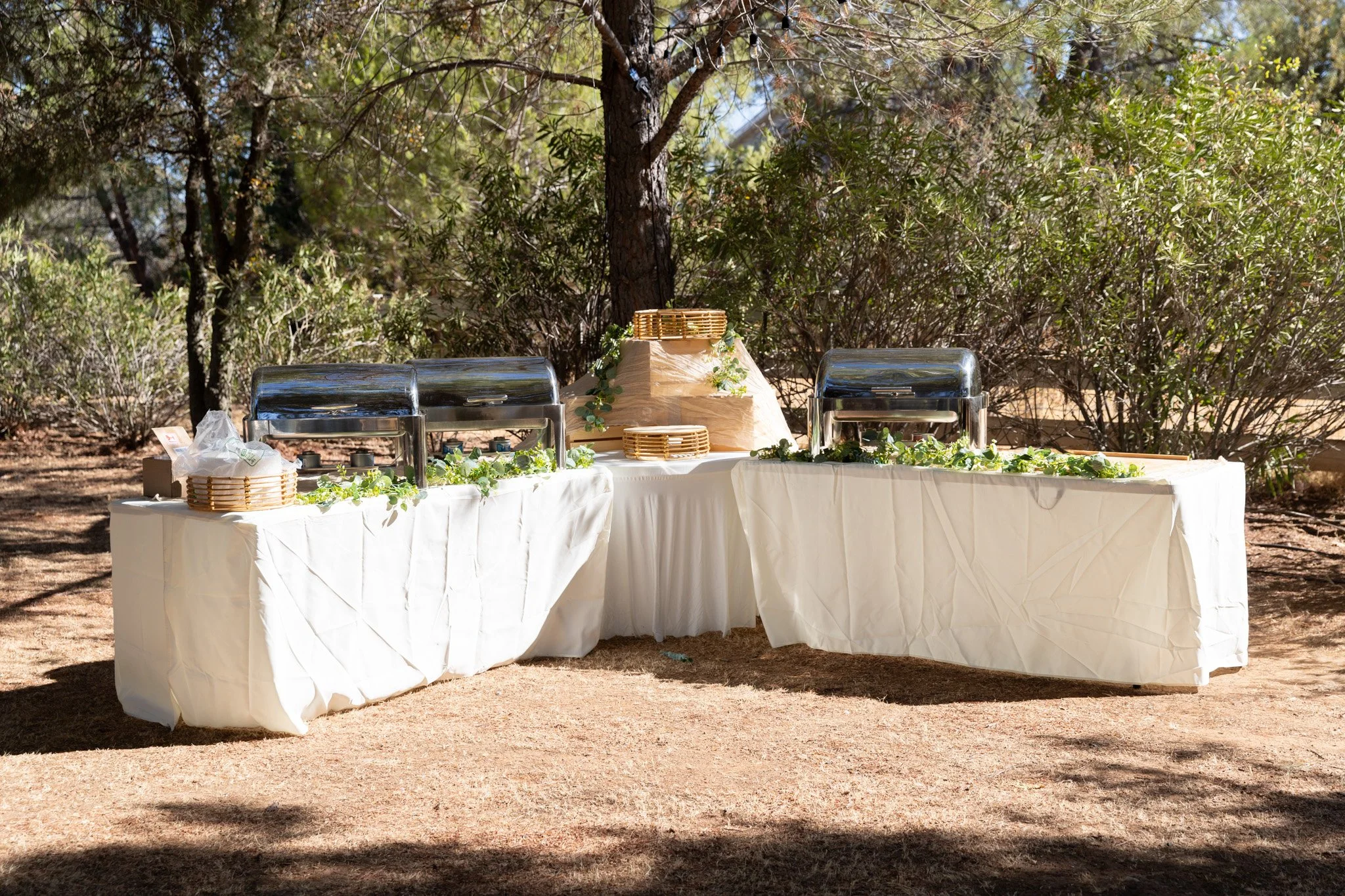 Outdoor buffet setup with white tablecloths, covered serving dishes, and greenery decorations, set against trees and shrubs.