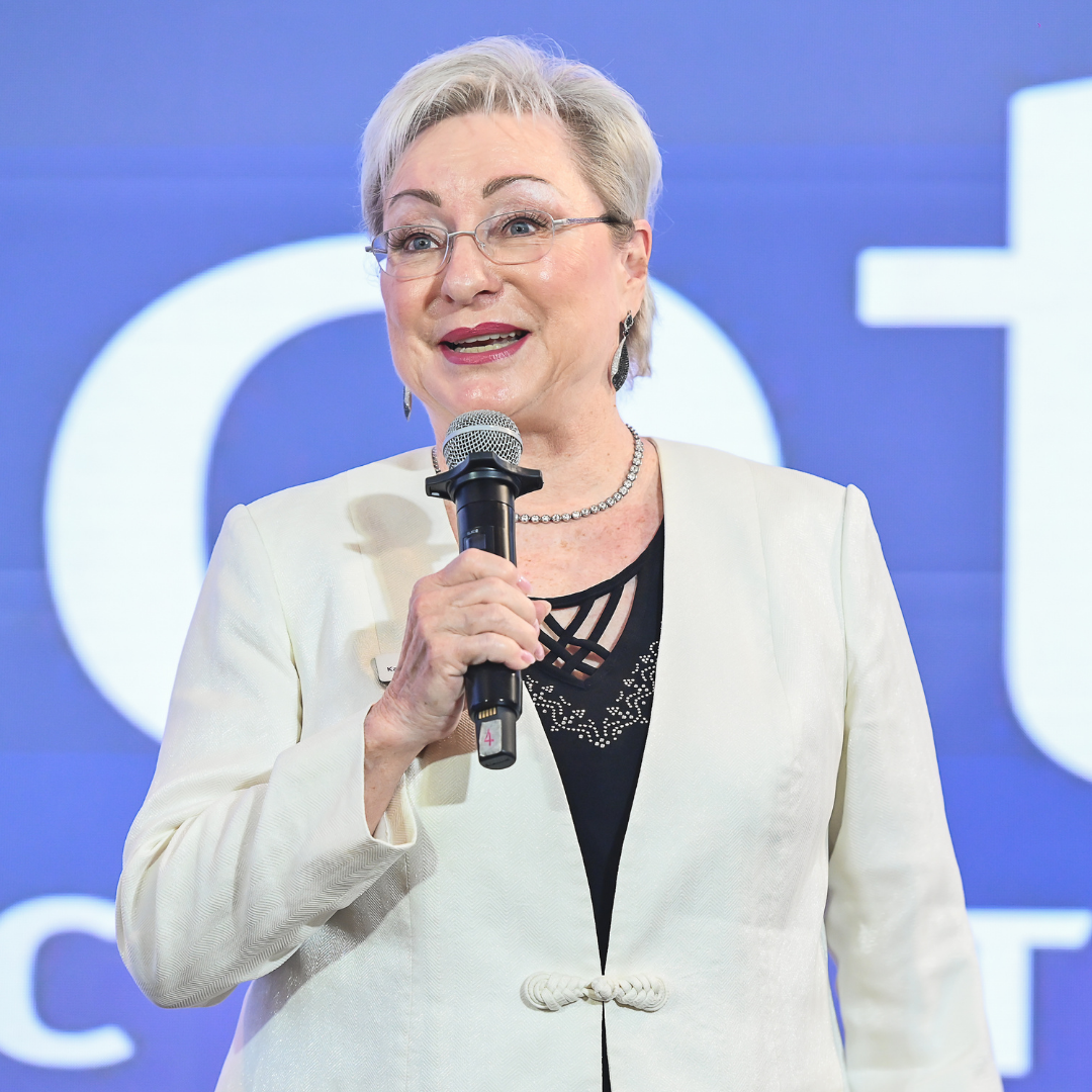 A woman with short gray hair, glasses, and earrings is speaking into a microphone at an event. She is wearing a white blazer over a black top with lace detailing.