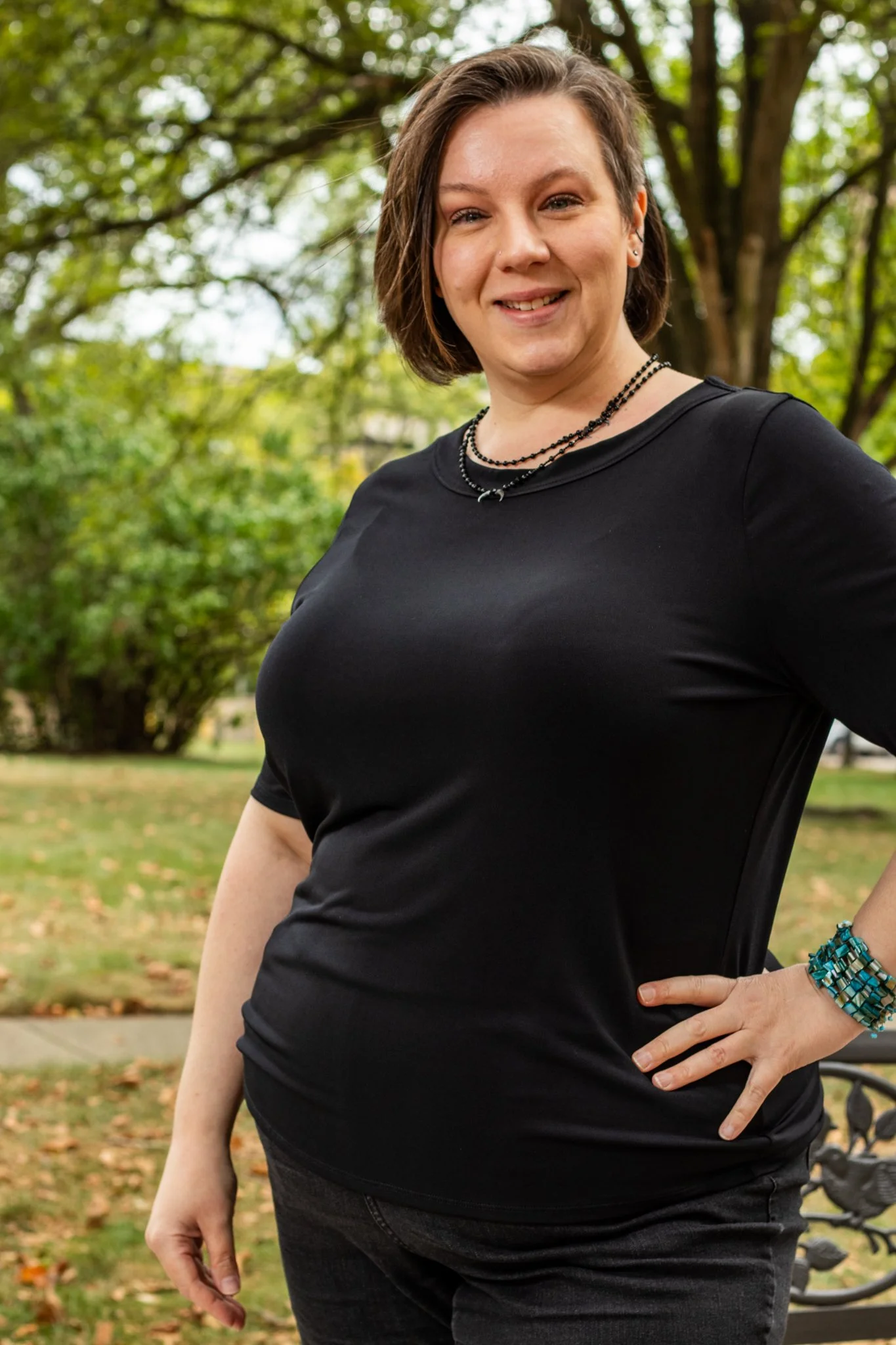 Dianne Goetsch, a woman with short brown hair wearing a black shirt, black jeans, and beaded jewelry standing outdoors in a park with green trees in the background.