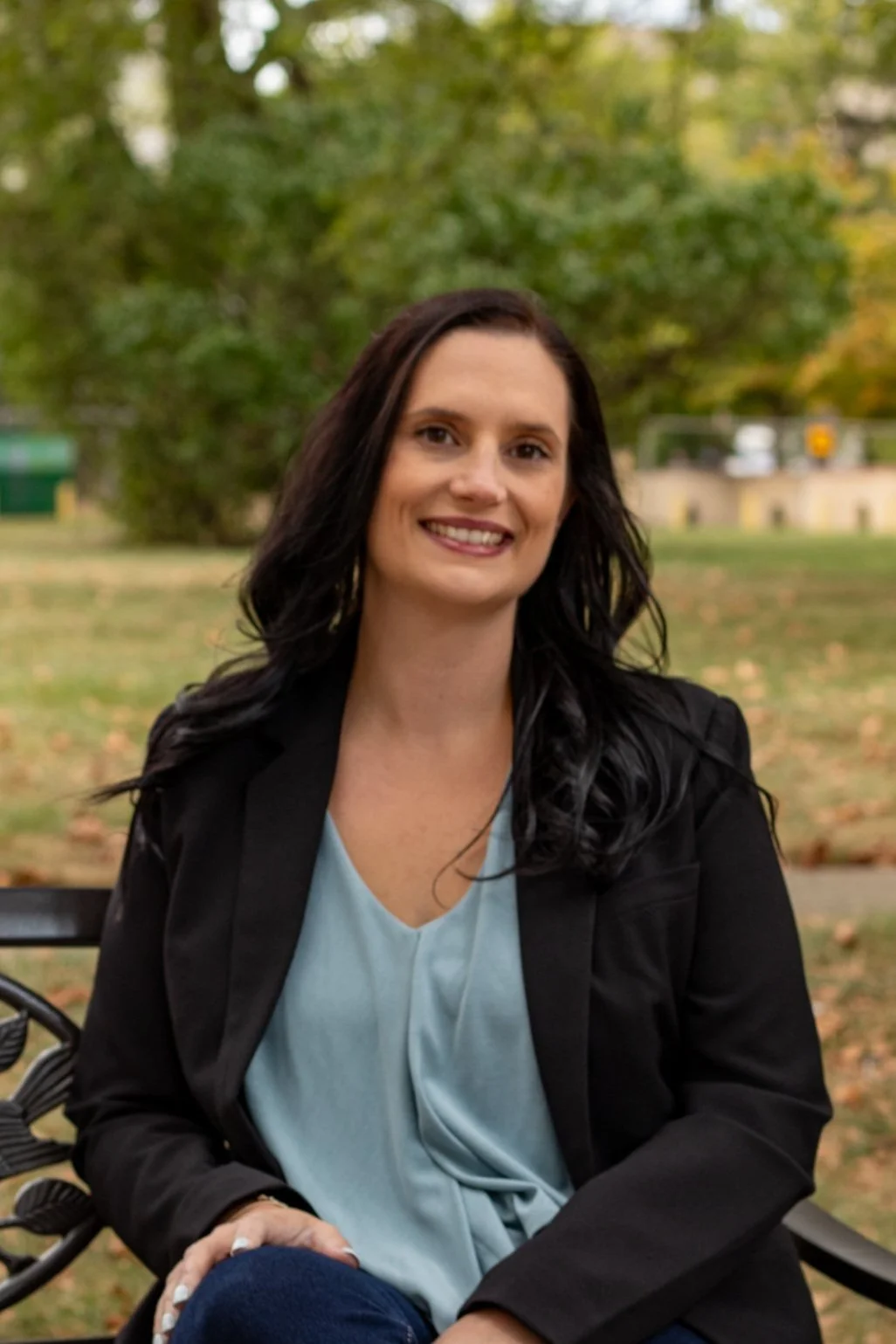 Jade Ellenbrook smiling, with long dark hair sitting on a park bench surrounded by green and yellow trees in autumn.