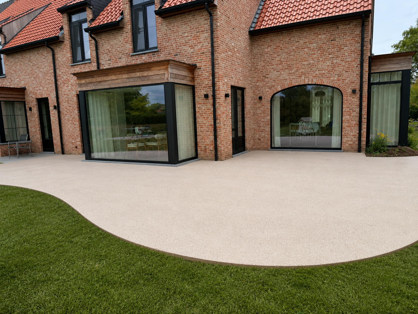 Moderne patio met lichtkleurig terras en tuin met groen gras, achter een huis met bakstenen buitenmuren en grote ramen.