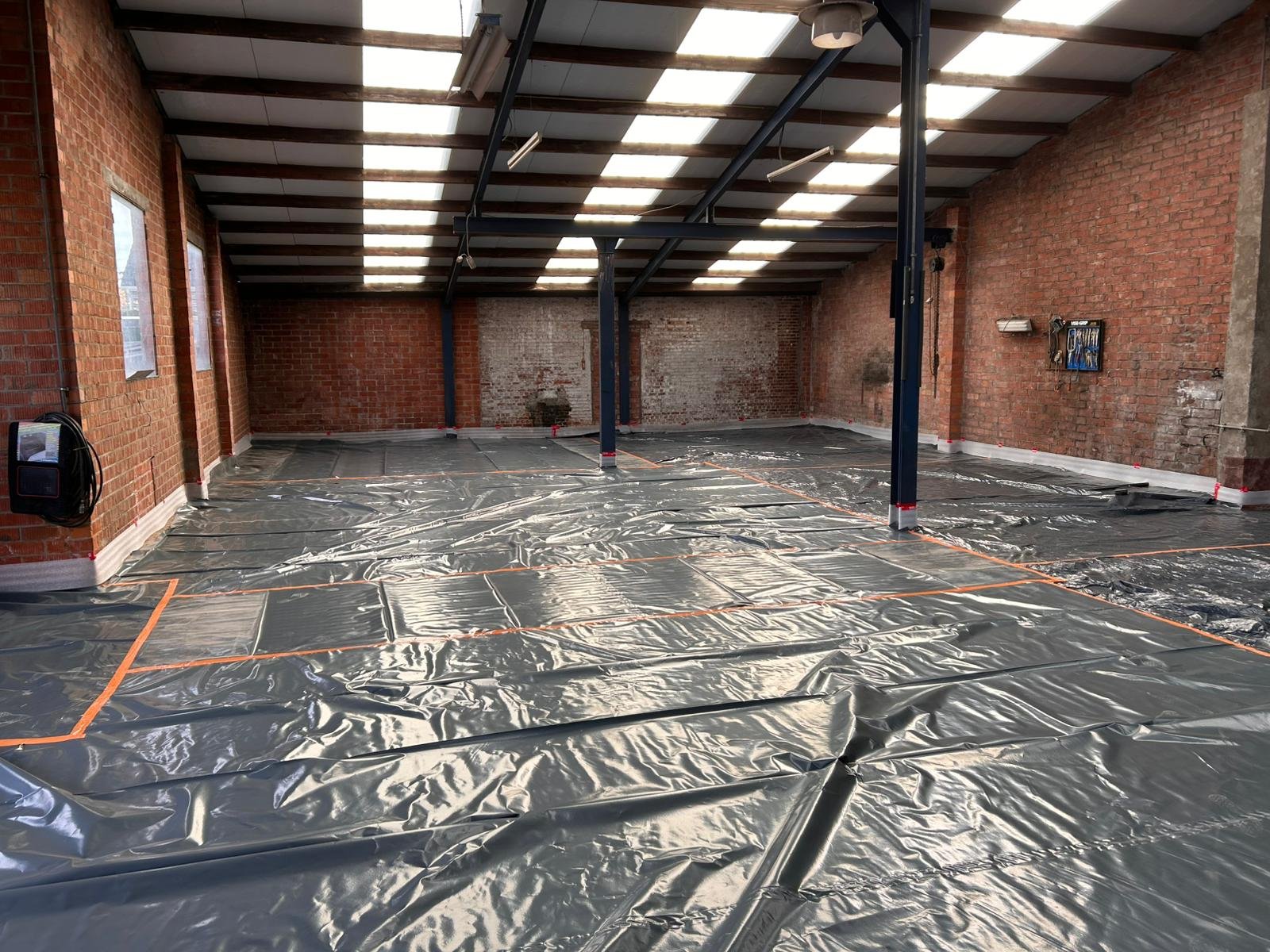 Binnenruimte met bakstenen muren en een metalen draagstructuur, vloer bedekt met reflecterend folie, waarschijnlijk voor bouw of renovatie.