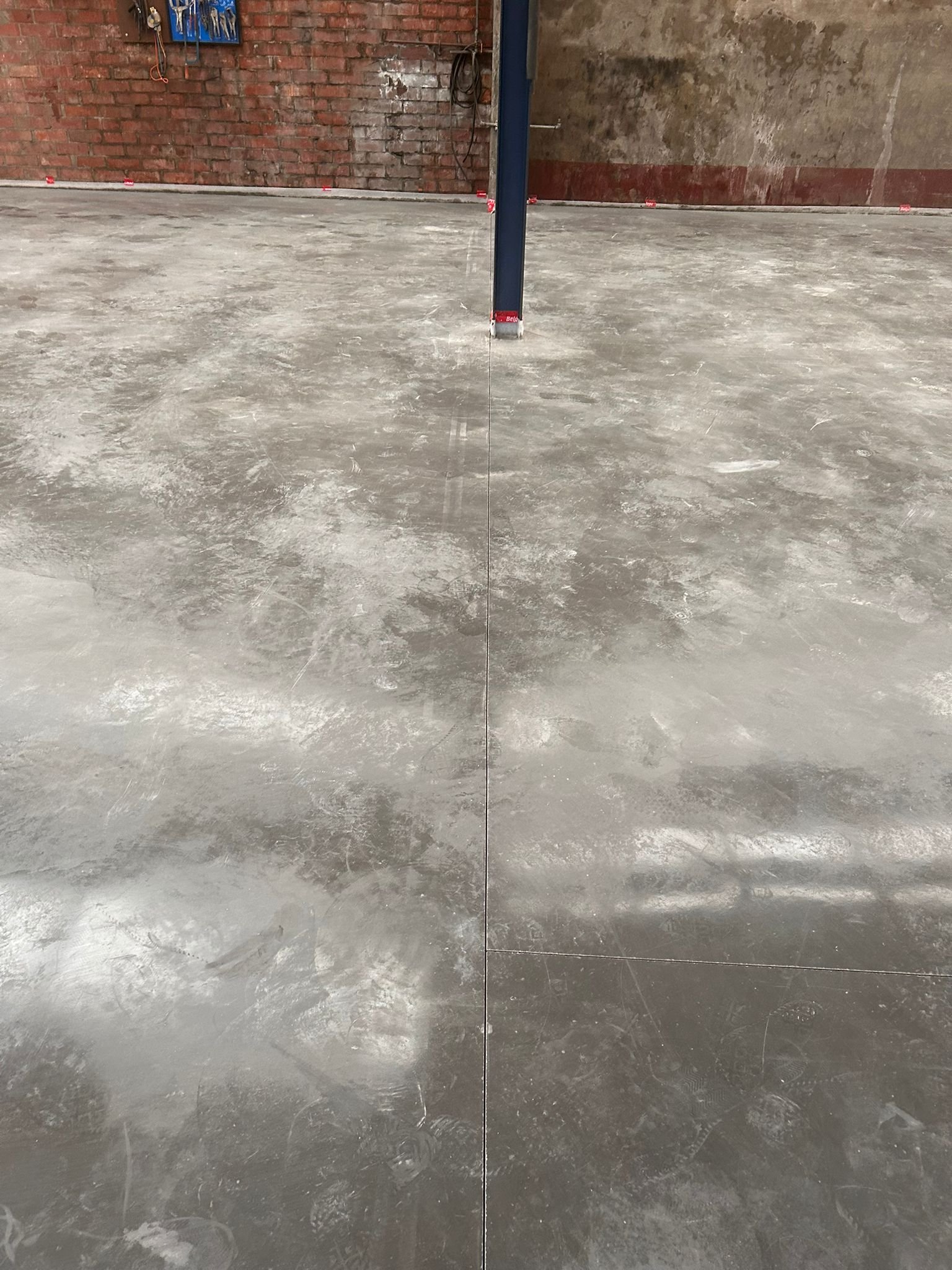 Foto van een betonvloer in een garage of werkplaats met een metalen steunpilaar in het midden en een bakstenen muur op de achtergrond.
