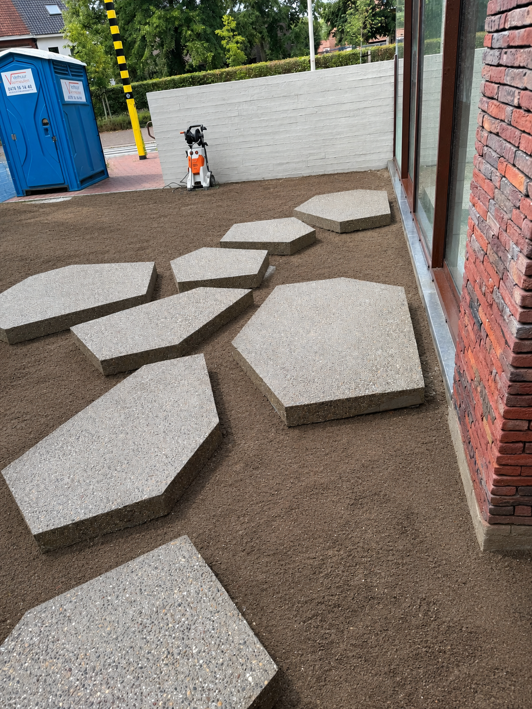 Bordenstenen worden geplaatst op een gronddekking in een tuin voor een pad, met een stenen muur en grote ramen naast de plek. Er is ook een tuin- of bouwmachine in de achtergrond.