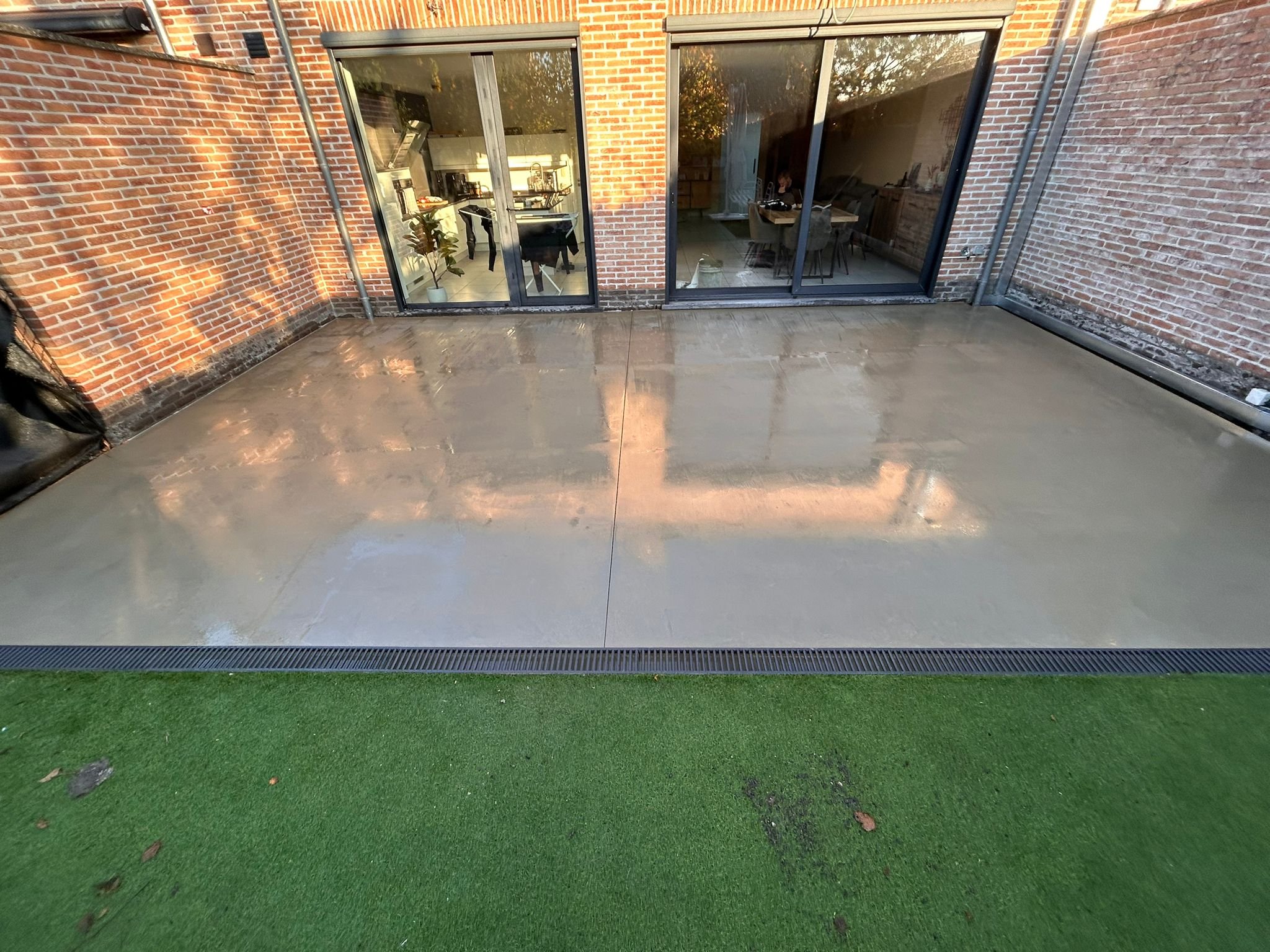 Buitenterras met betegelde vloer en uitzicht op een woonkamer met grote glazen deuren en een tafel met stoelen binnen.