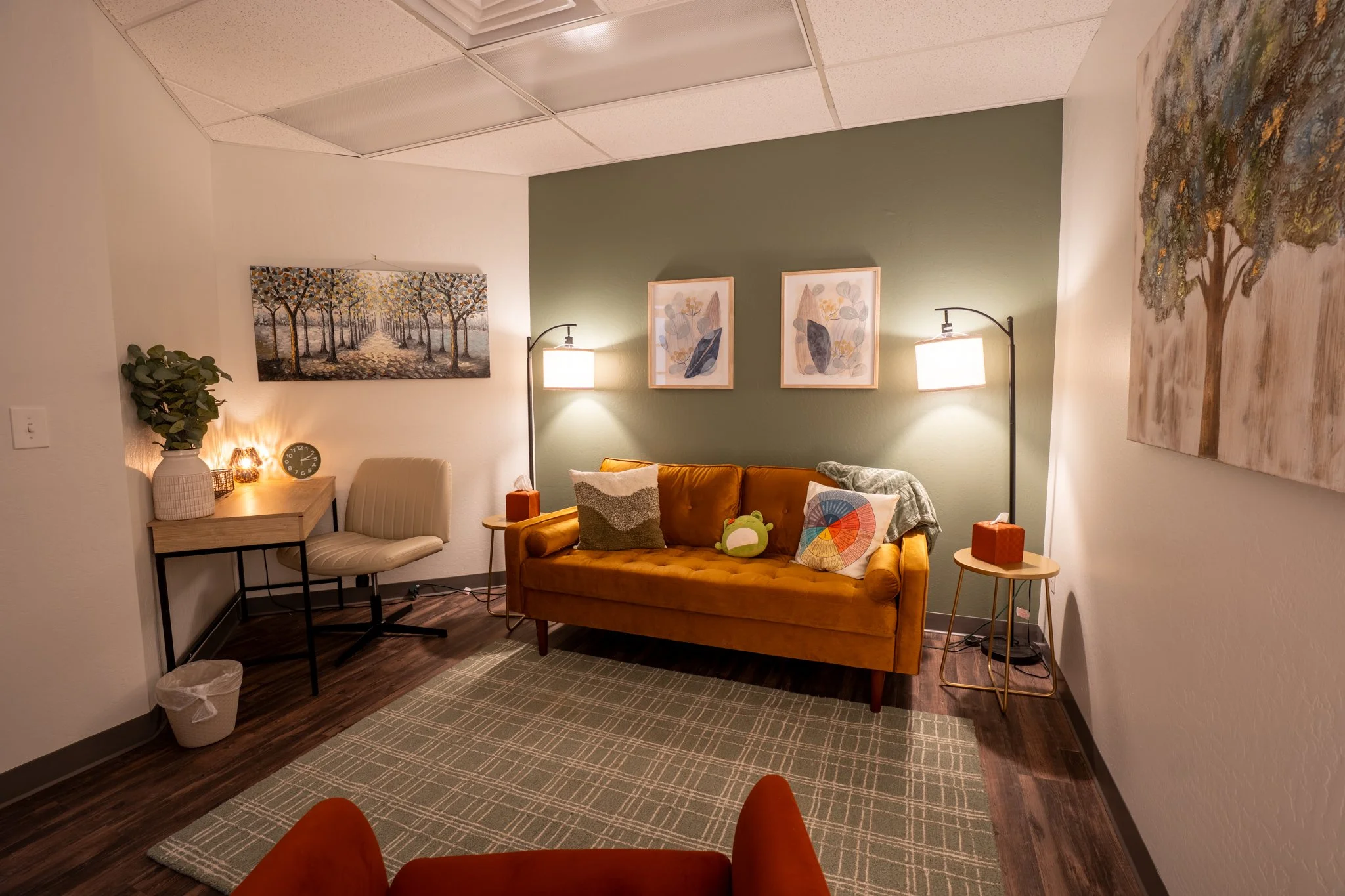 A cozy room with a mustard-colored couch, a white chair, and two standing lamps with white shades. There are framed art pieces on the wall, including a landscape of trees and abstract art, and a potted plant on a wooden side table. The room has a patterned carpet and dark wooden flooring.