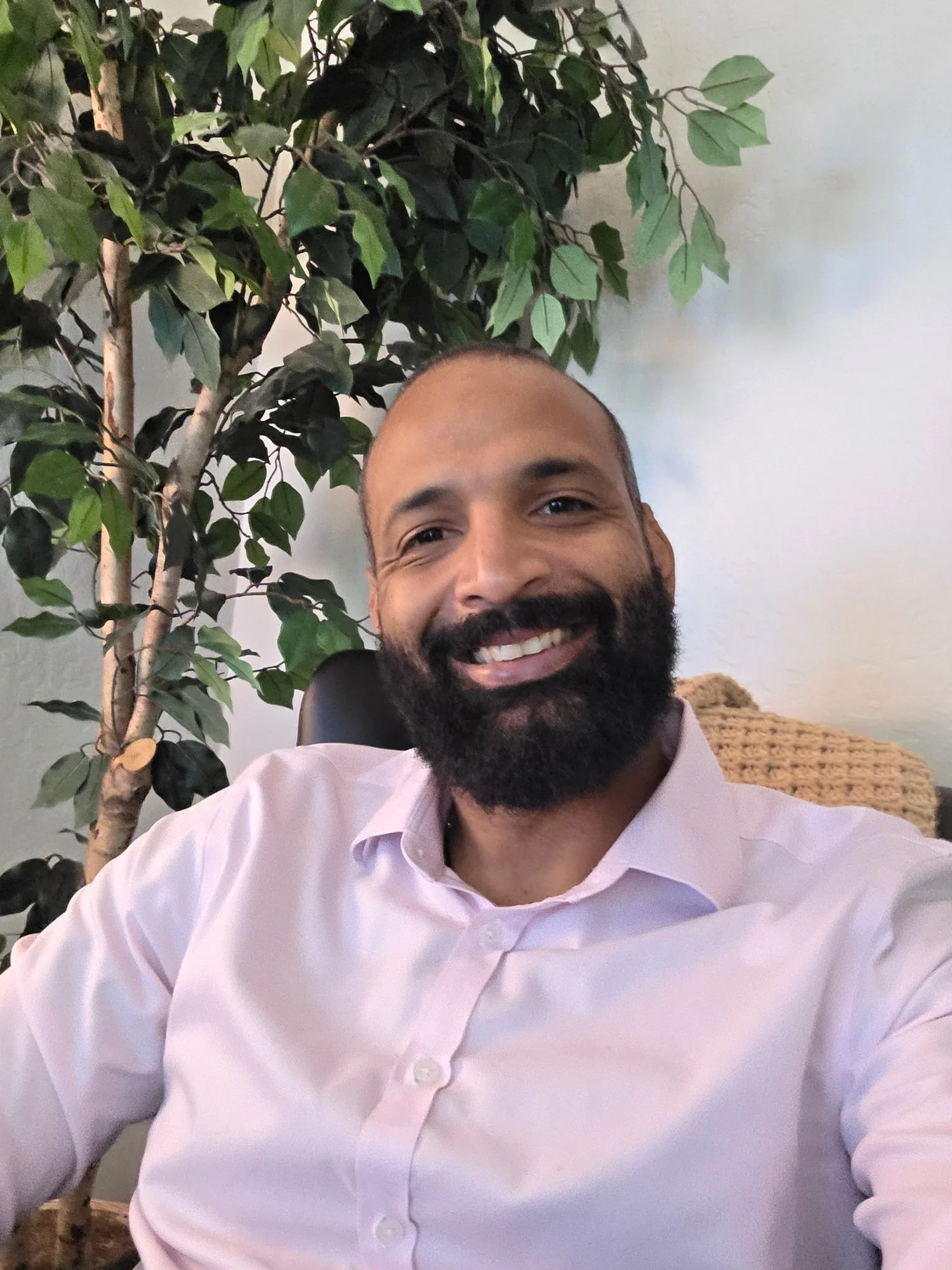 Smiling man with a beard and mustache wearing a light pink shirt, sitting in front of a large green leafy plant, indoor setting.