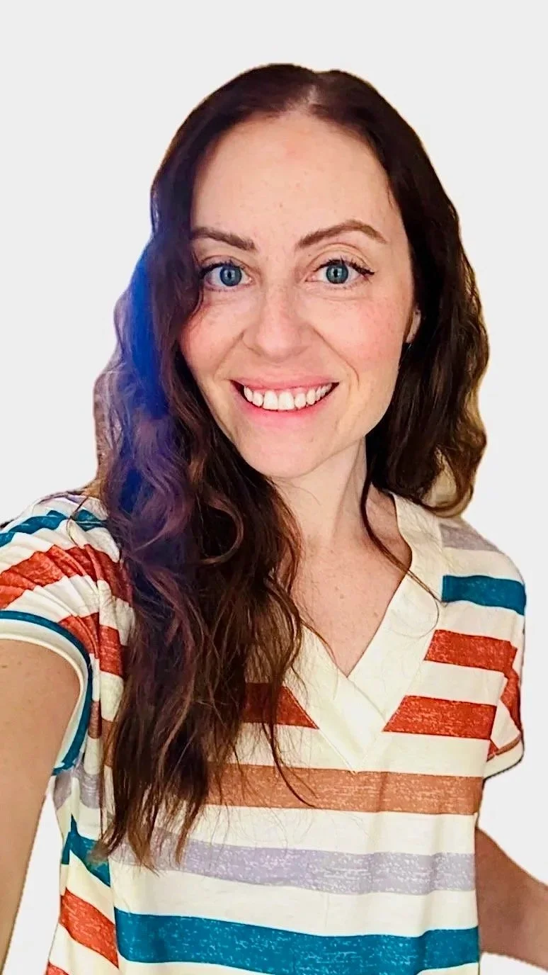 A young woman with long wavy brown hair and blue eyes smiling, wearing a colorful striped t-shirt with red, blue, lavender, and beige horizontal stripes.