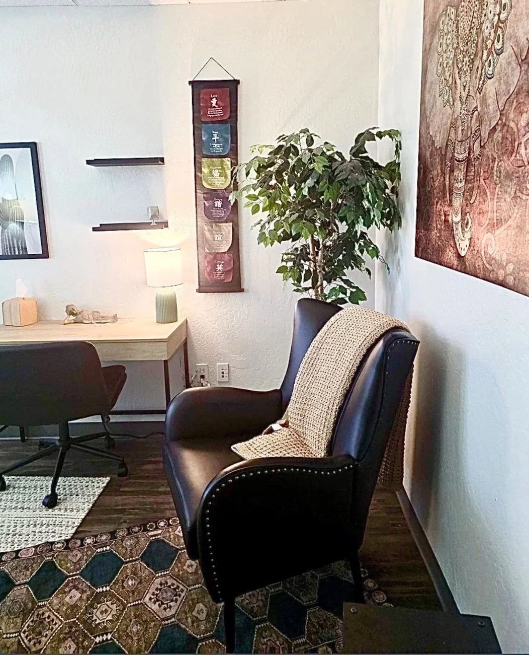 A cozy corner of a room with a black leather armchair with nailhead trim and a beige knitted blanket draped over the back, a tall green potted plant, a wooden desk with a small lamp and tissue box, a black office chair, wall decorations including a vertical colorful tapestry and a framed picture, and a patterned area rug.