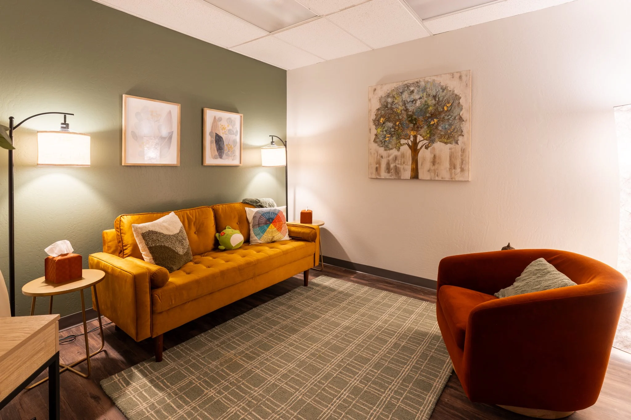 Therapy office with green and white walls, featuring a yellow sofa with pillows, a matching yellow armchair, a side table with tissue box, framed wall art, and a rug with a checkered pattern.