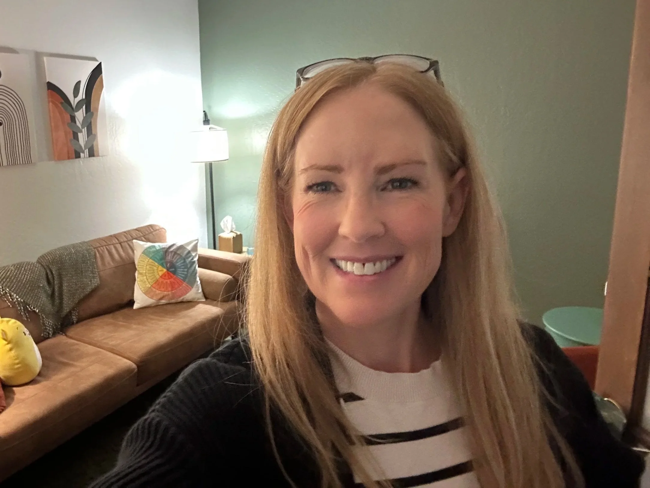 Close-up of a smiling woman with blonde hair and glasses resting on her head, in a warmly lit room with a green wall, artwork, a lampshade, a beige couch with colorful pillows, and a small green side table.