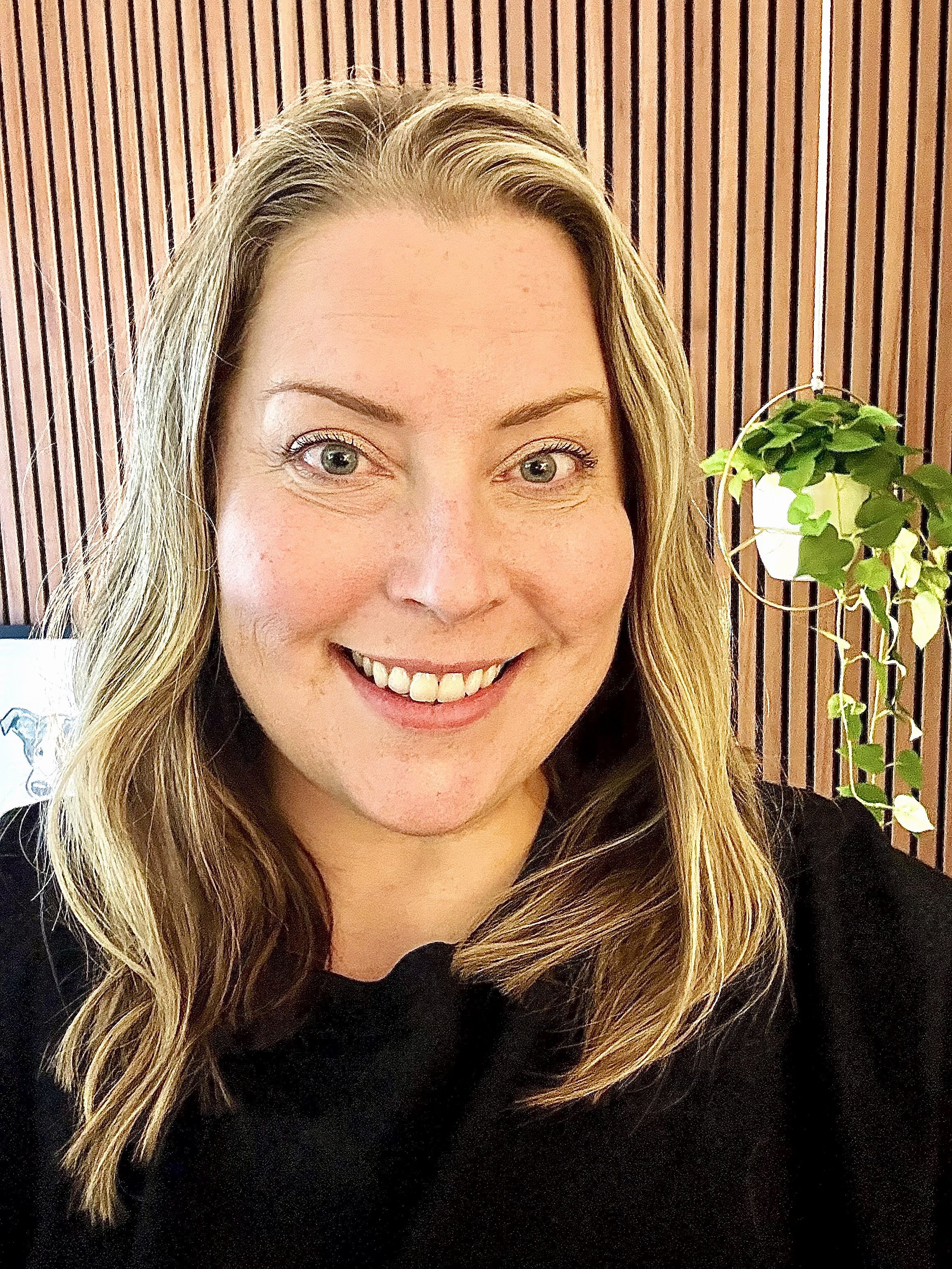 A woman with shoulder length wavy blonde hair, blue eyes, and a bright smile, wearing a black top, with a wood slatted panel and green hanging plant in the background.
