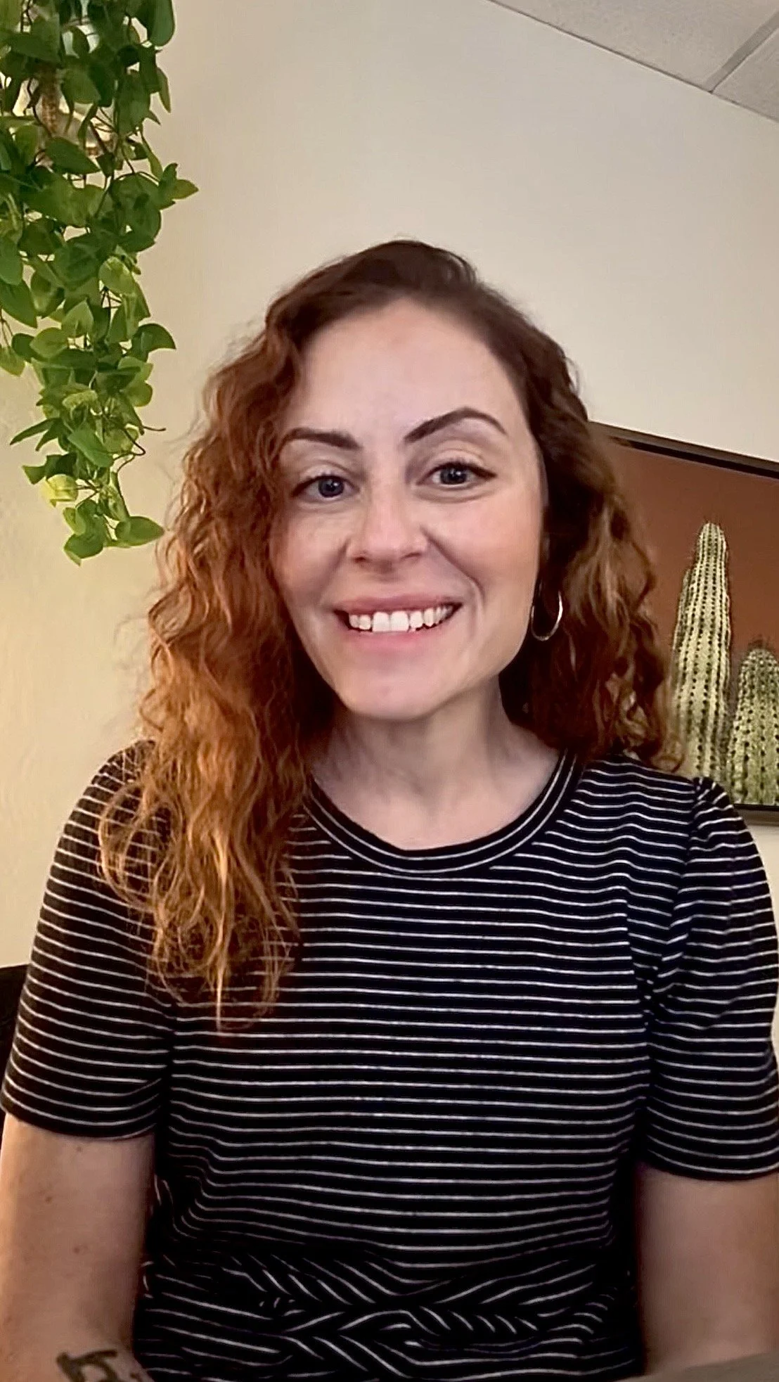 A young woman with long wavy brown hair and blue eyes smiling, wearing a colorful striped t-shirt with red, blue, lavender, and beige horizontal stripes.