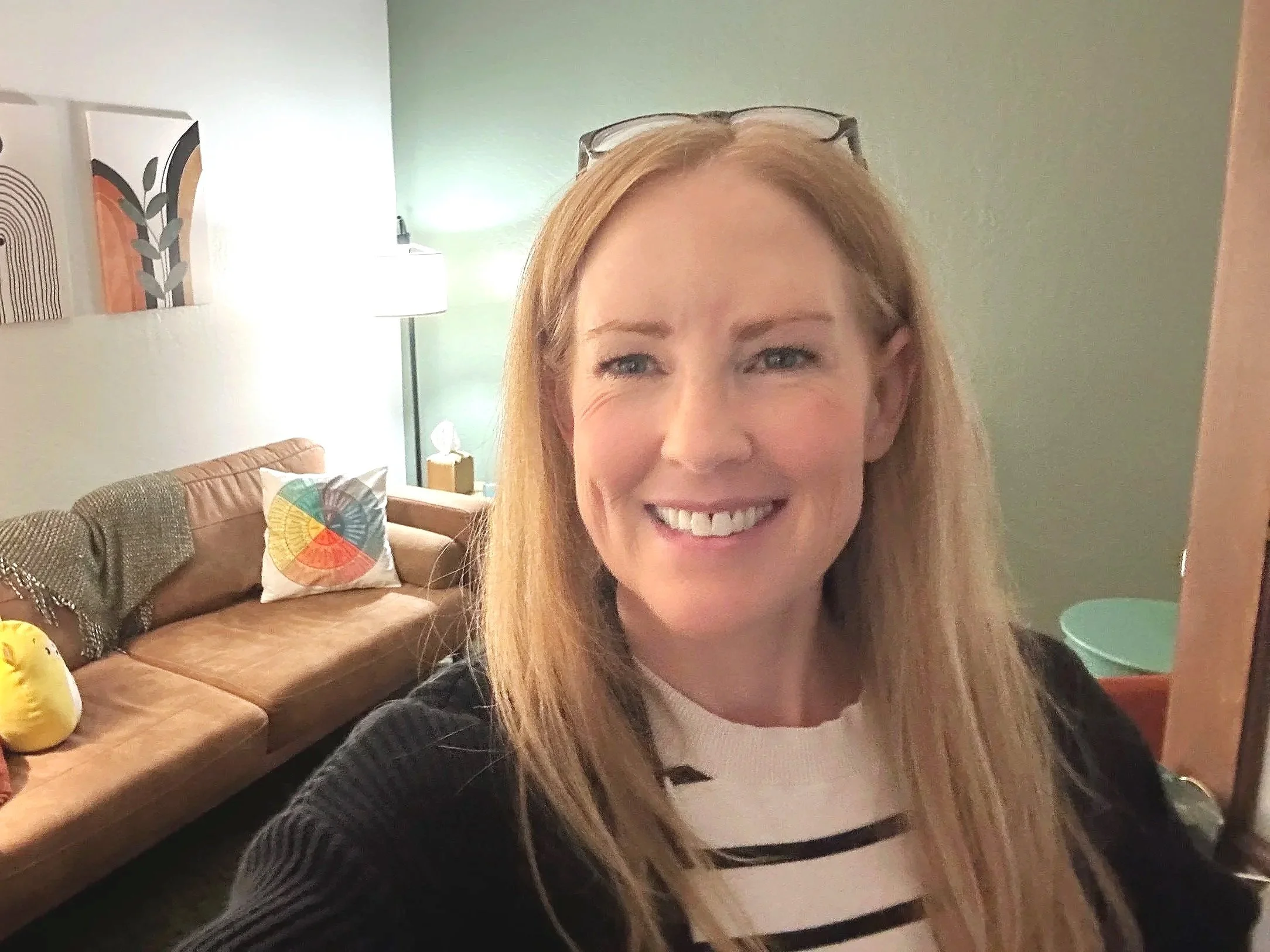 Close-up of a smiling woman with blonde hair and glasses resting on her head, in a warmly lit room with a green wall, artwork, a lampshade, a beige couch with colorful pillows, and a small green side table.