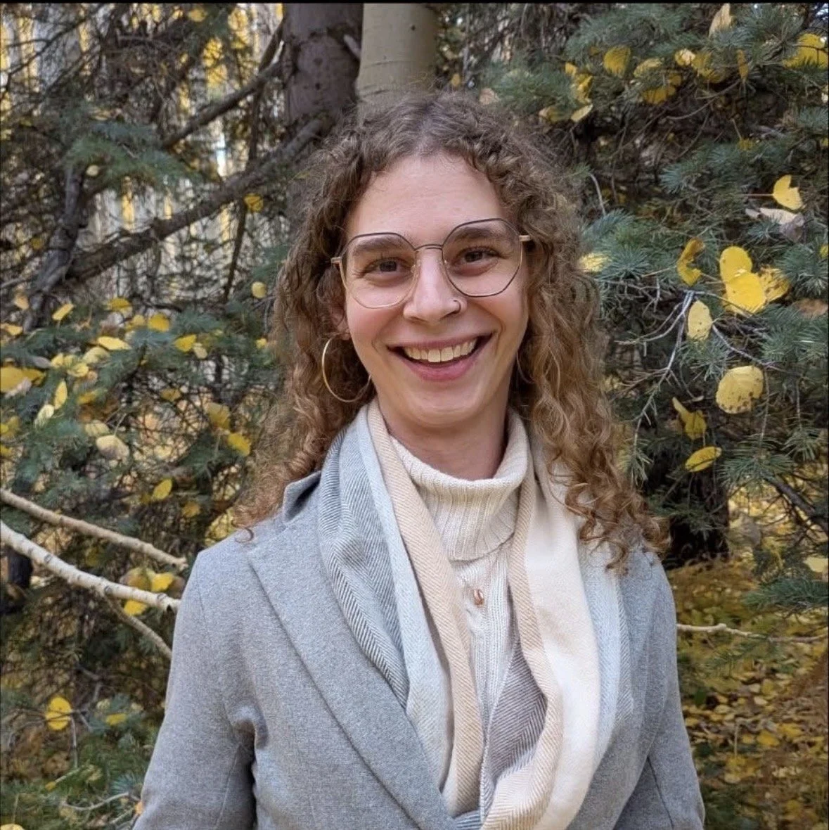A woman with curly hair, glasses, and a nose ring smiling outdoors in front of a pine tree with yellow leaves.