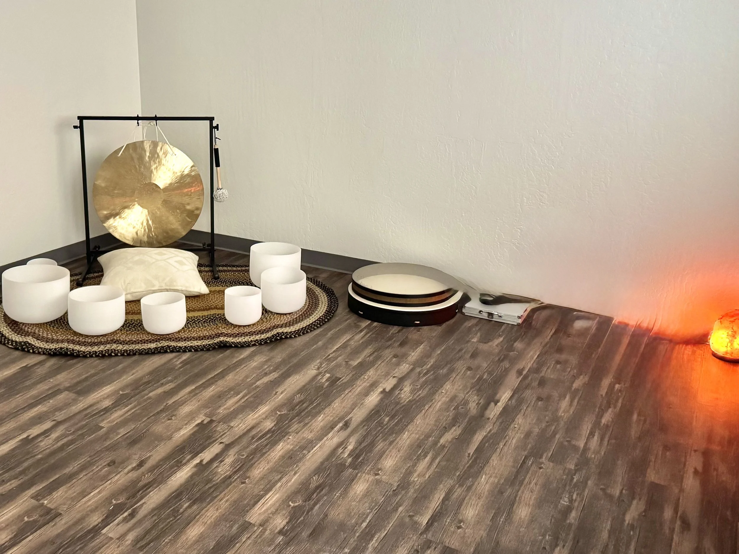 A meditation space with a large golden gong on a black metal stand, a white cushion, white singing bowls, and a lava lamp on the floor, with the light from the lava lamp casting a warm glow.
