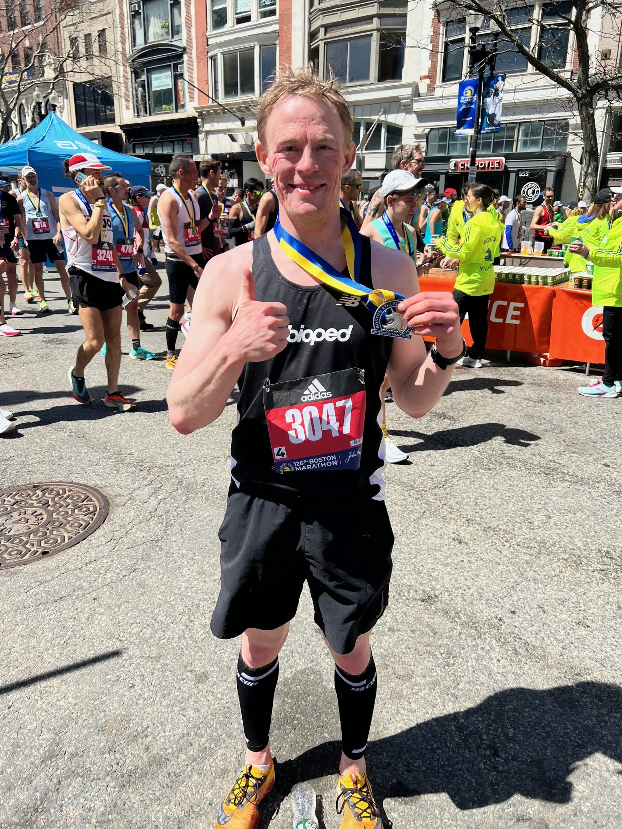 A male marathon runner with bib number 3047, smiling and giving a thumbs up after finishing a race at the Boston Marathon, surrounded by other runners and volunteers.