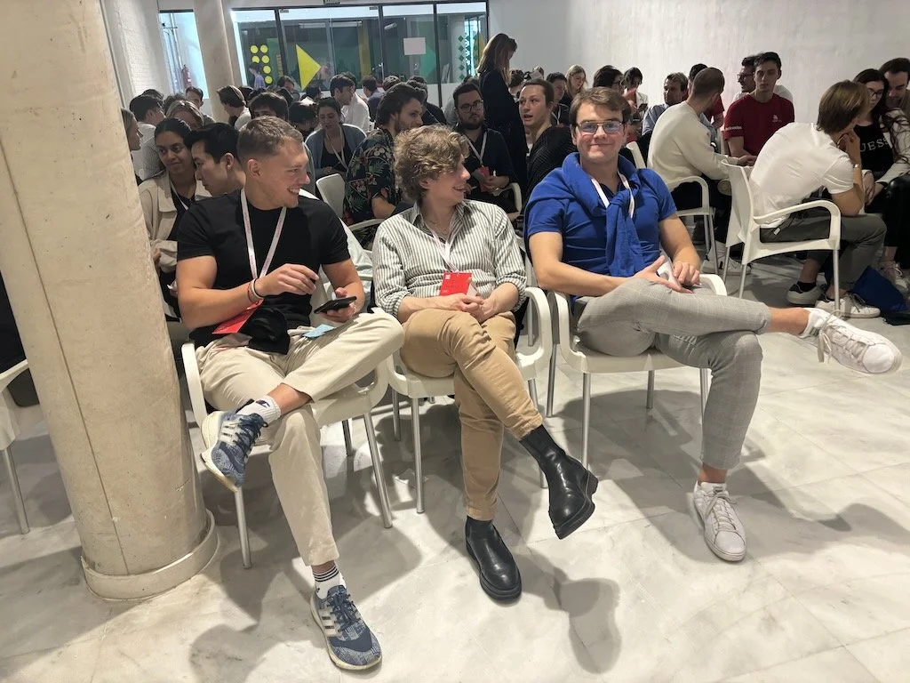 Group of people sitting in chairs at an indoor event, engaged in conversation and using smartphones, with a glass door and colorful wall decor in the background.