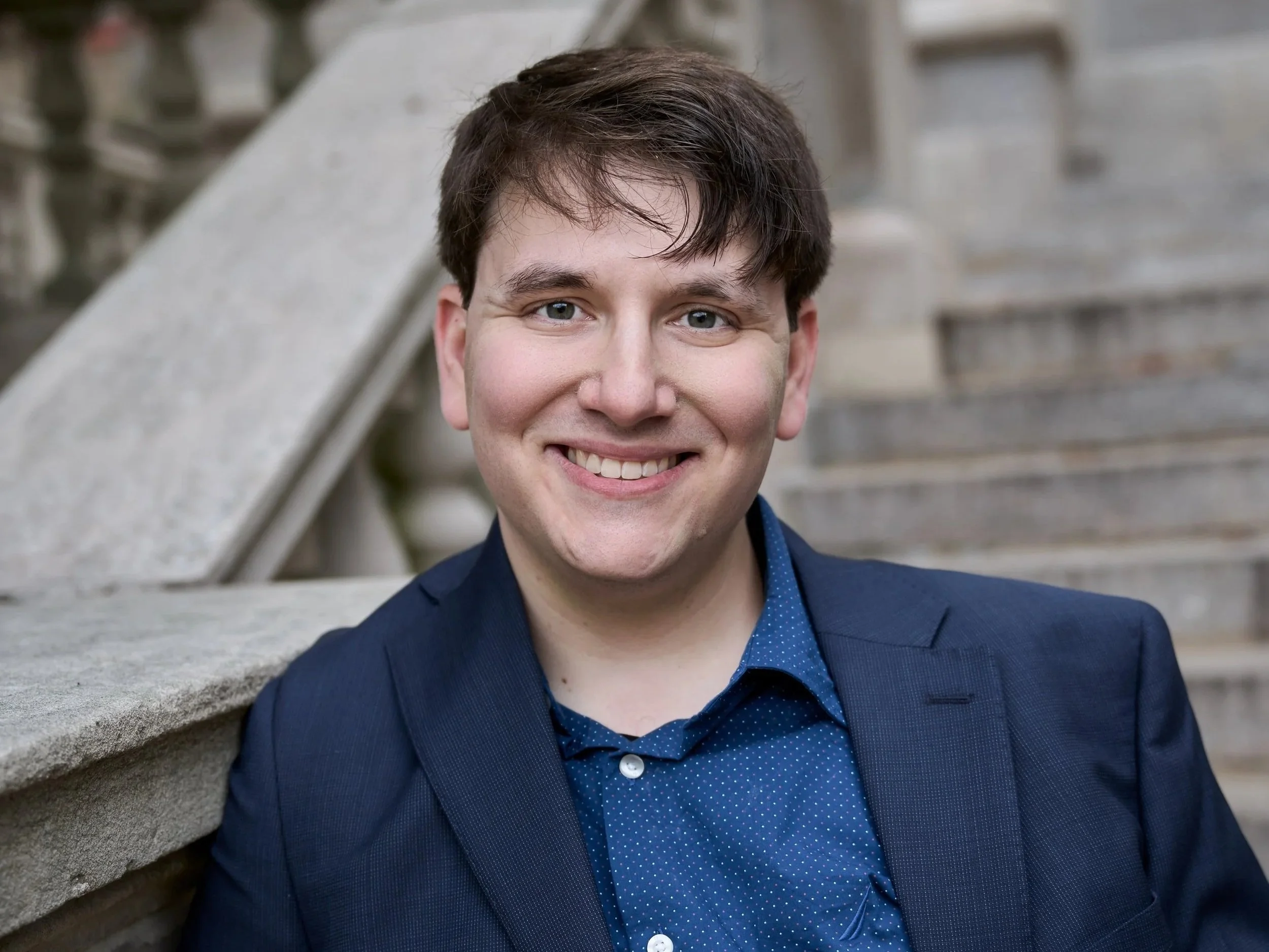 A young man with short brown hair, blue eyes, and light skin, smiling in front of stone stairs and railing, wearing a navy blue suit and a blue polka-dot shirt.
