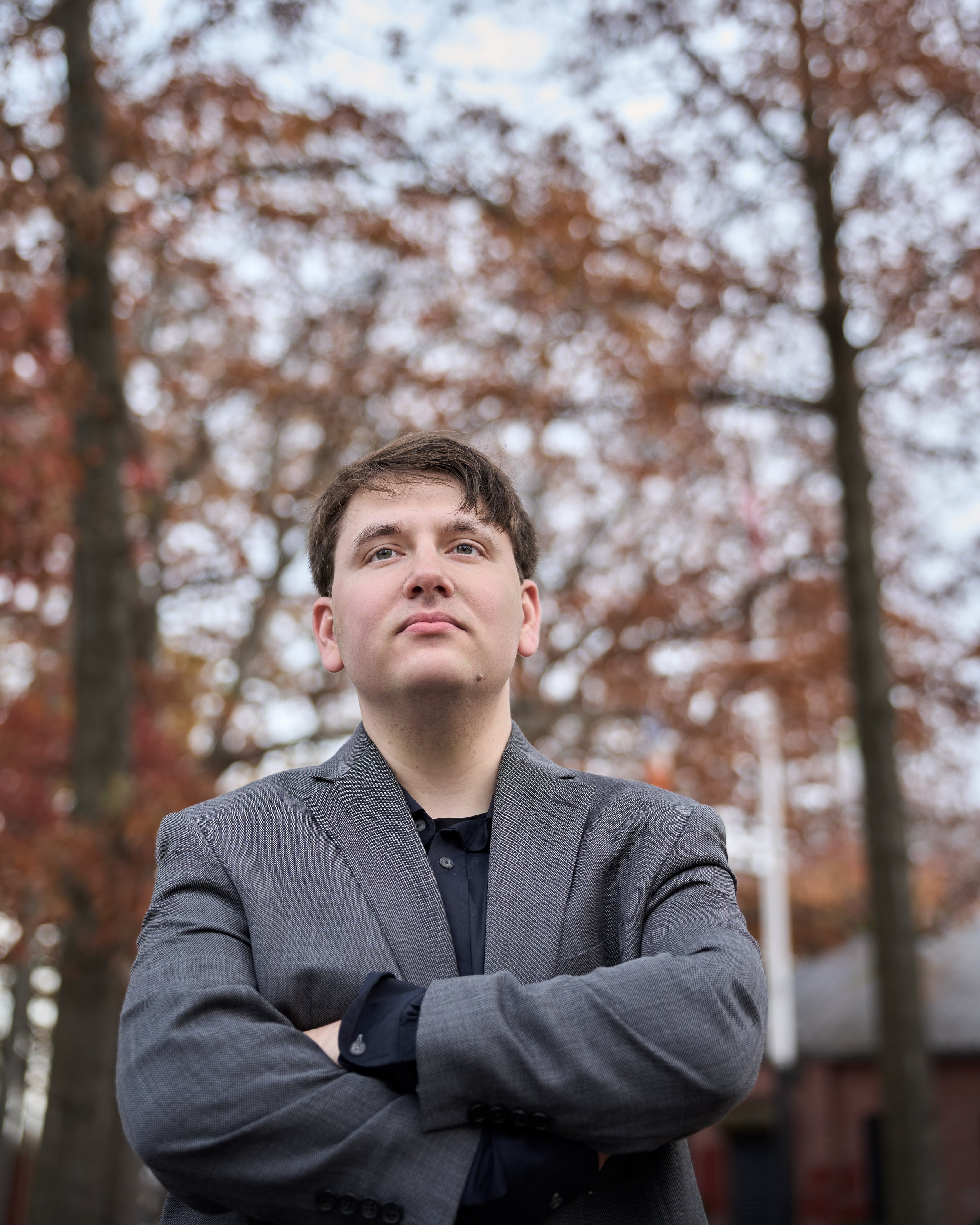 A man in a gray blazer with crossed arms, looking confident outdoors with autumn colored trees in the background.