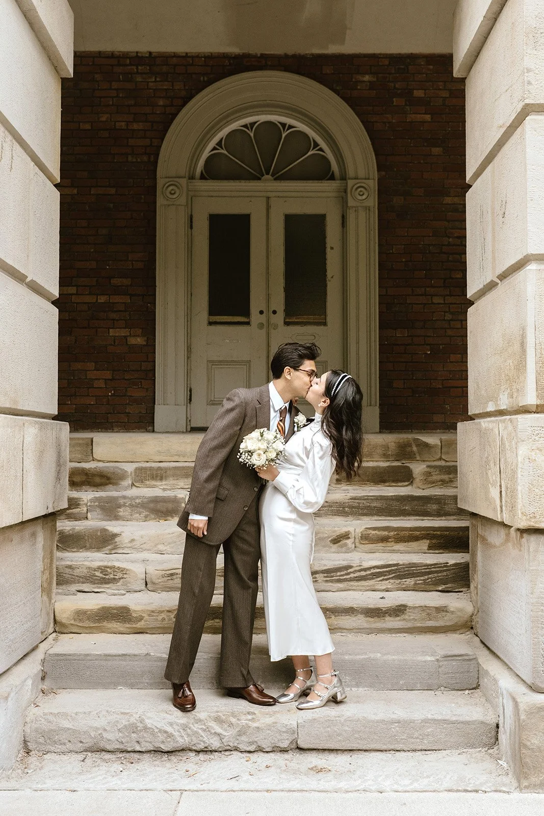 A bride and groom sharing a kiss on a stone staircase outside toronto city hall with a large arched wooden door