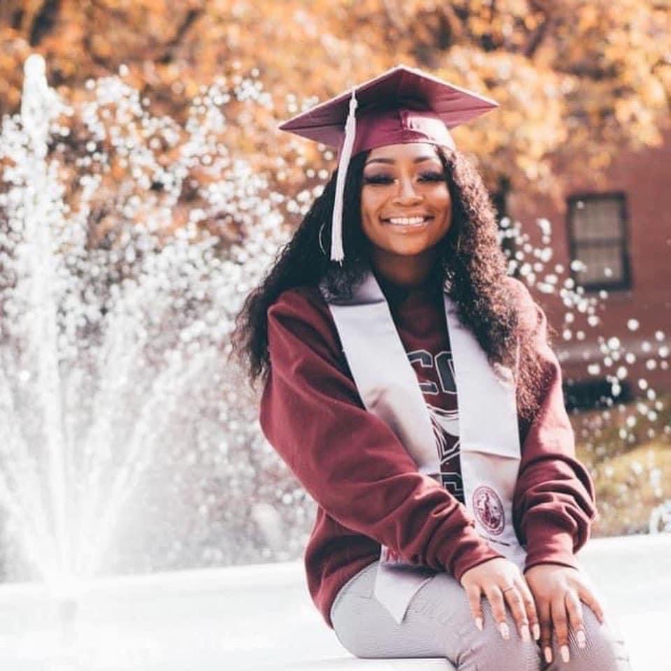 A young woman in a graduation cap and gown smiling outdoors, with autumn trees and a fountain in the background.
