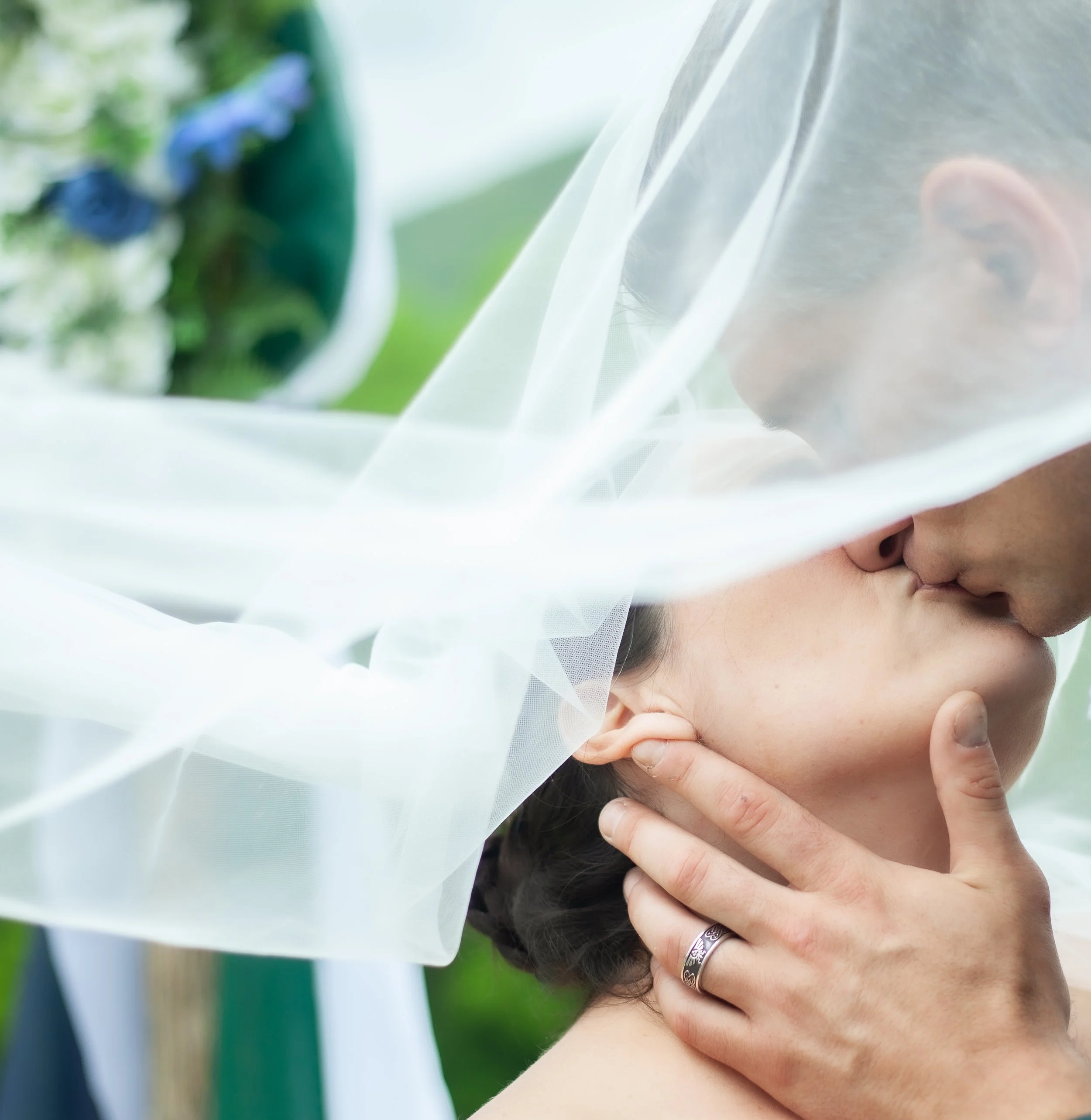 A bride and groom share a kiss, partially obscured by a white bridal veil, with a bouquet of blue flowers in the background.