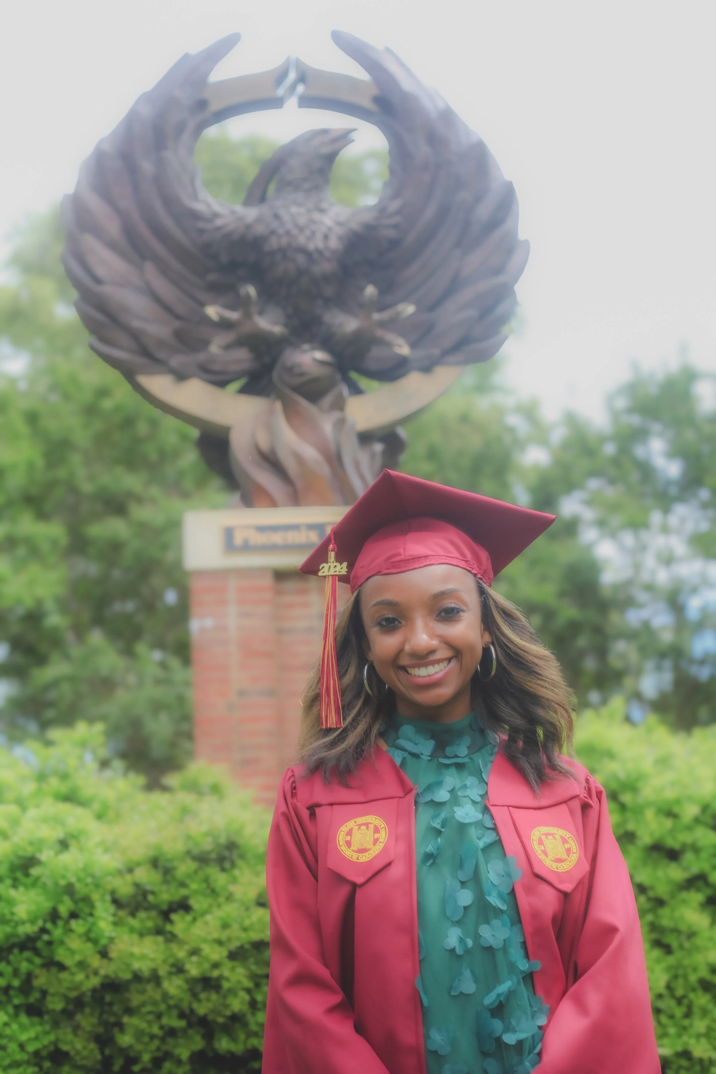 A young woman in a maroon graduation gown and cap, smiling outdoors with green trees and a fountain featuring an eagle statue in the background.