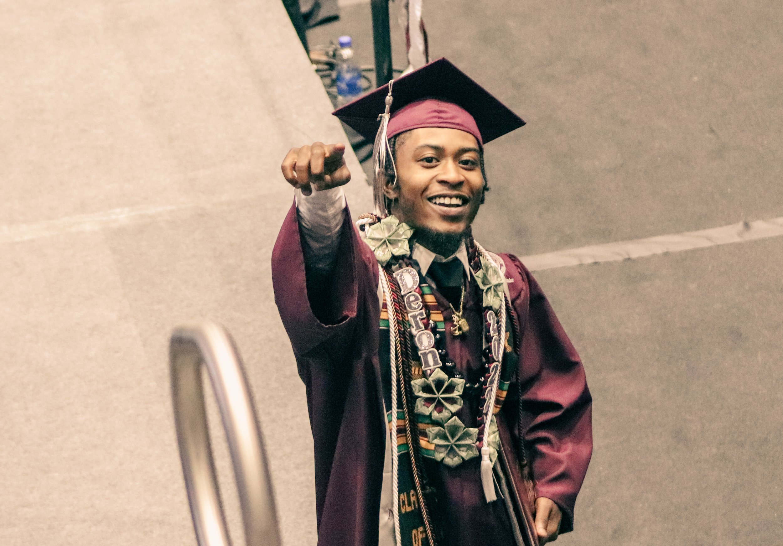 Young man in maroon graduation gown and cap, smiling and pointing forward, with leis and medals around his neck.