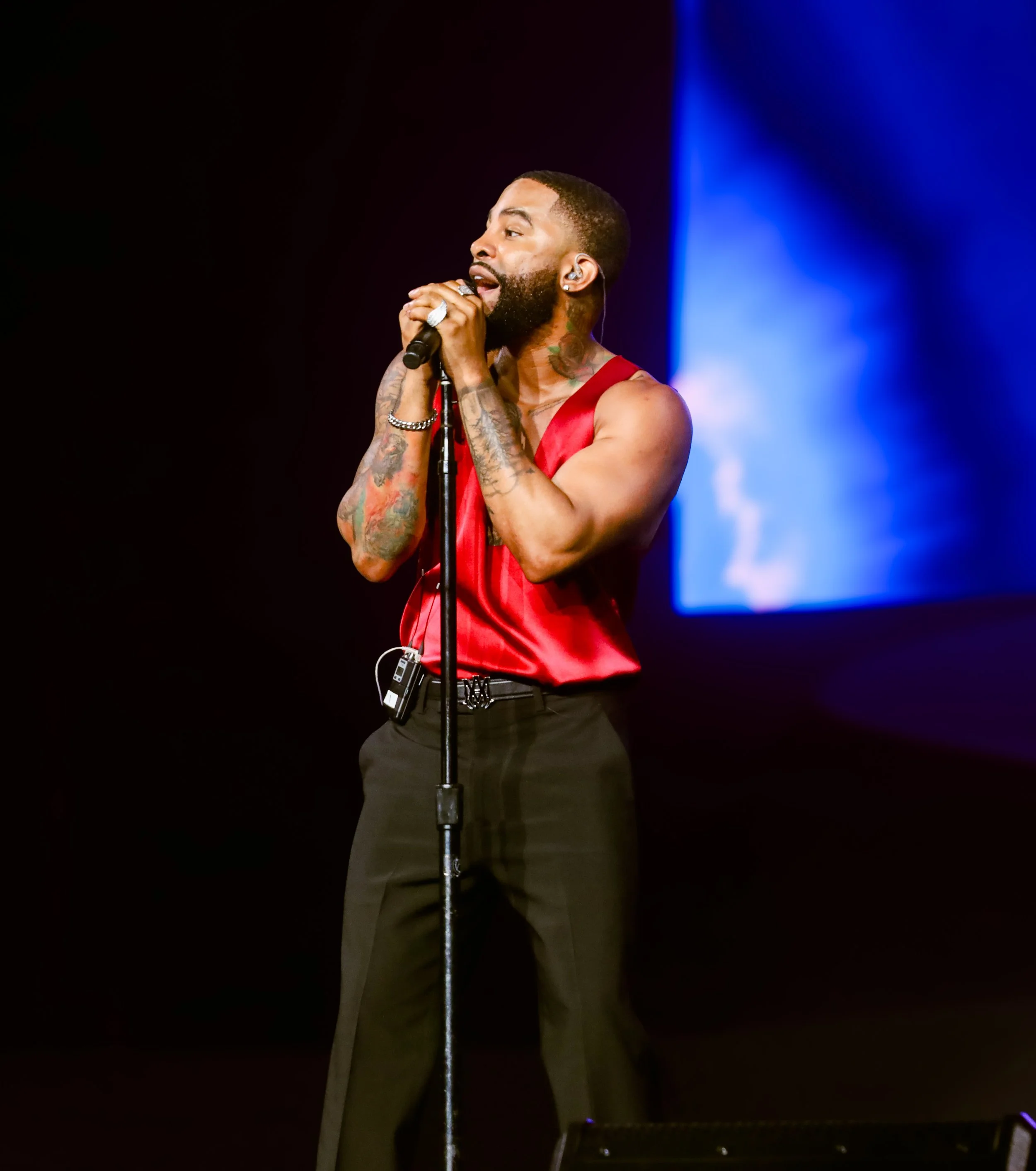 A man with tattoos singing into a microphone on stage, wearing a red satin sleeveless shirt and black pants, with a blue screen in the background.