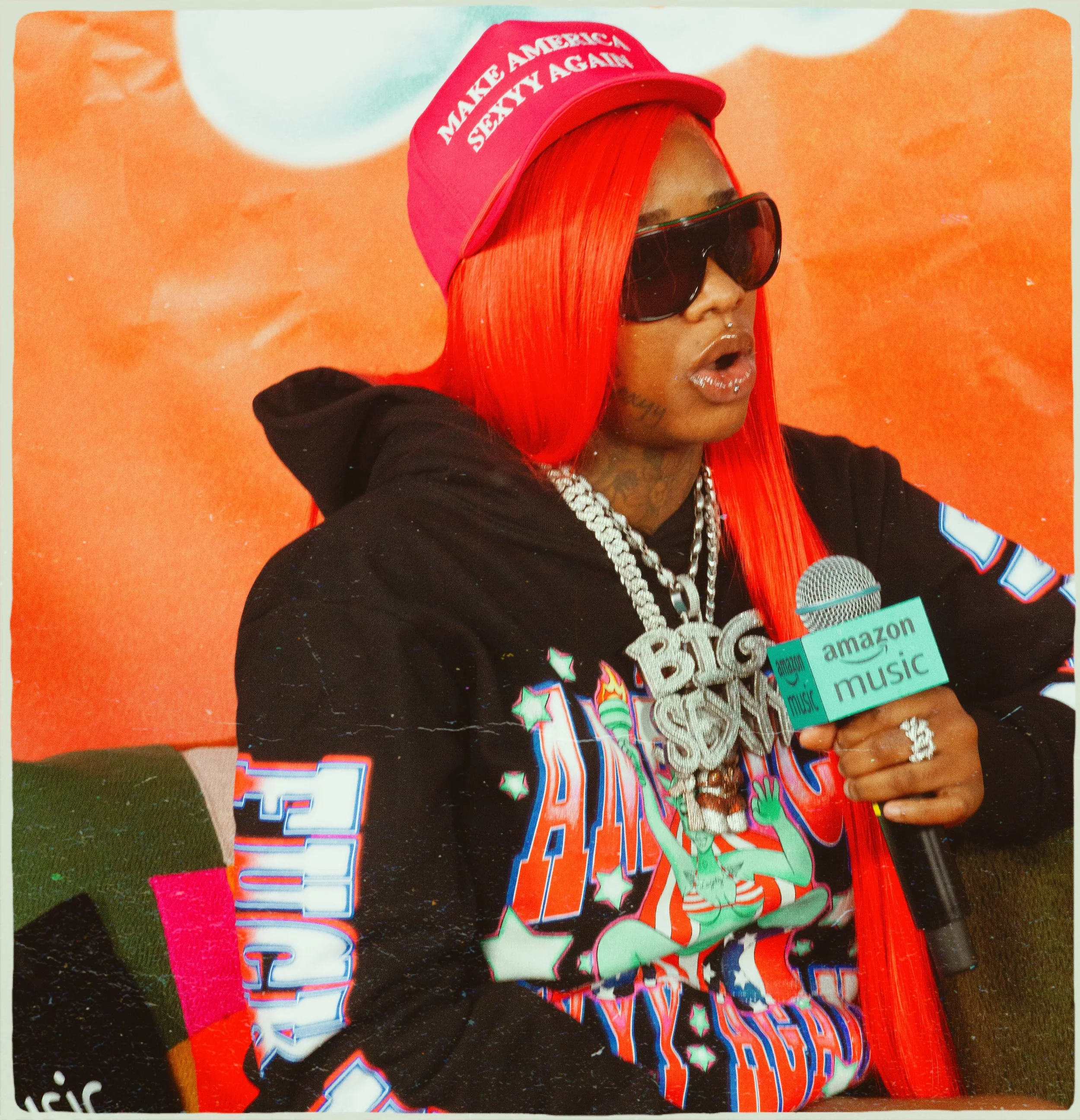A woman with long, bright red hair wearing a red hat that says "Make America Seventy Again," large sunglasses, a black hoodie, and oversized jewelry, holding a microphone that says "Amazon Music," in front of an orange background.