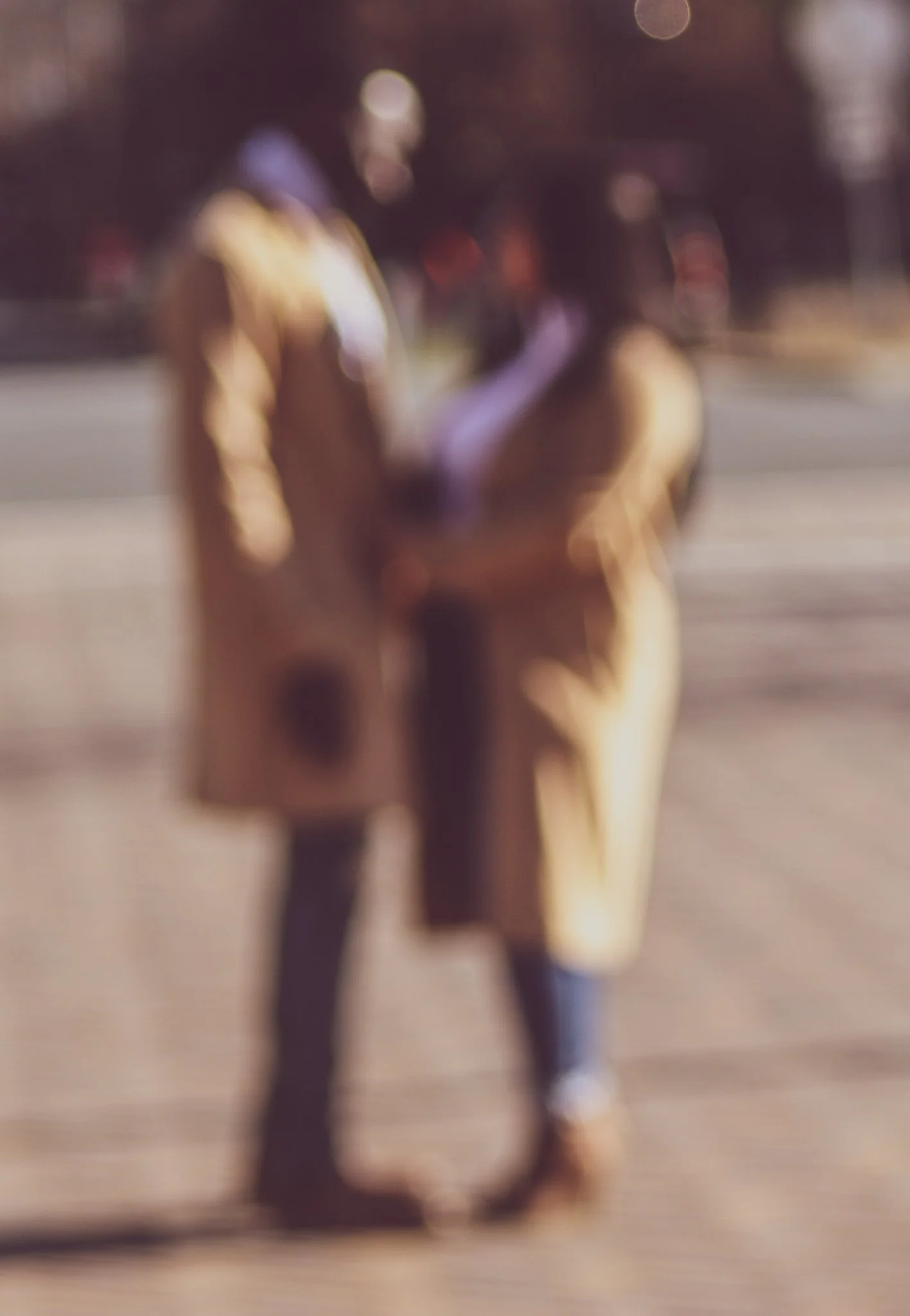Two people are standing outdoors in warm clothing, possibly during autumn or winter, engaging in conversation. The background shows a street with some blurred lights and trees.
