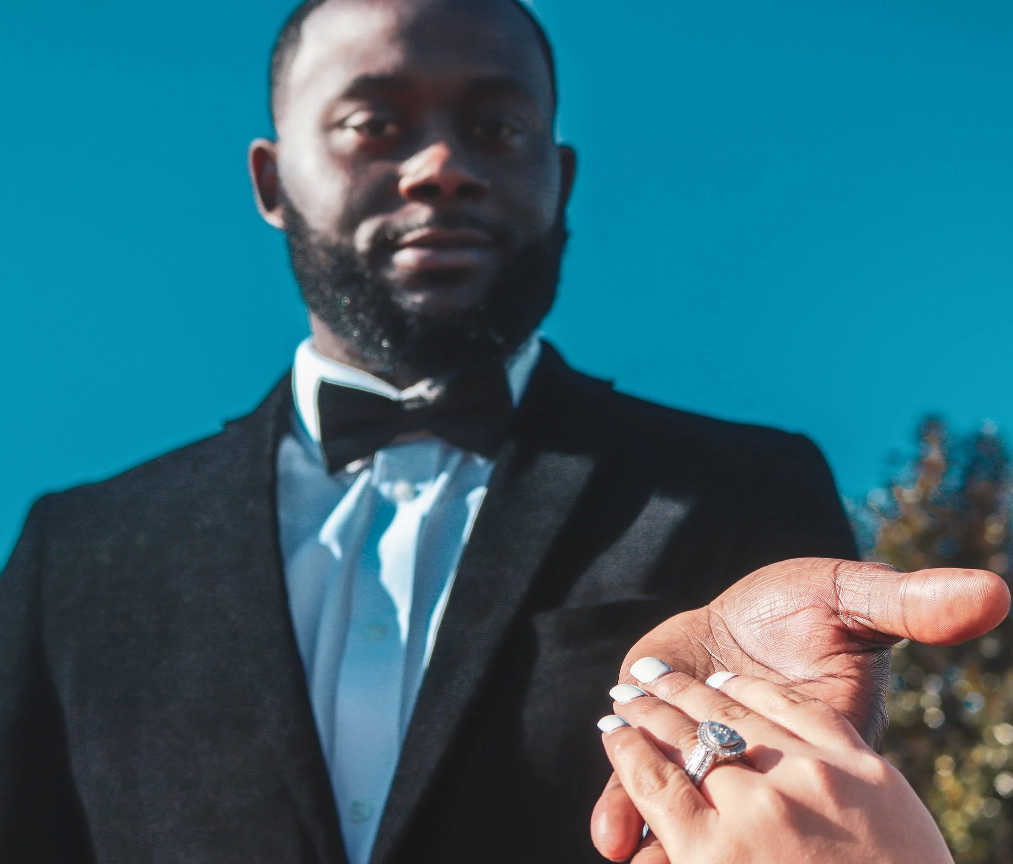 A man in a formal tuxedo with a bow tie, looking at the camera, with a woman showing her hand with wedding rings, against a background of a blue sky and some trees.