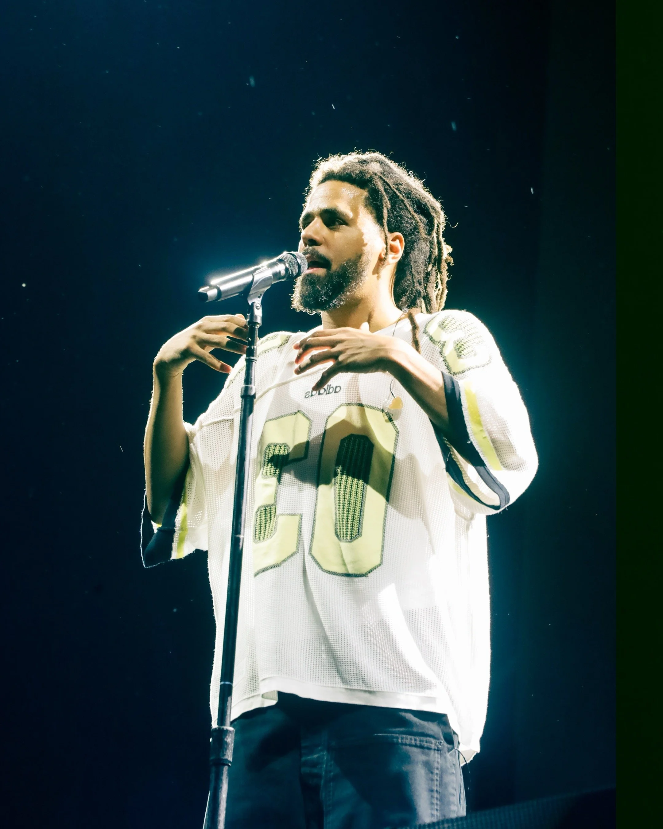 A man with dreadlocks and a beard performing on stage with a microphone, wearing a white football jersey with the number 20.