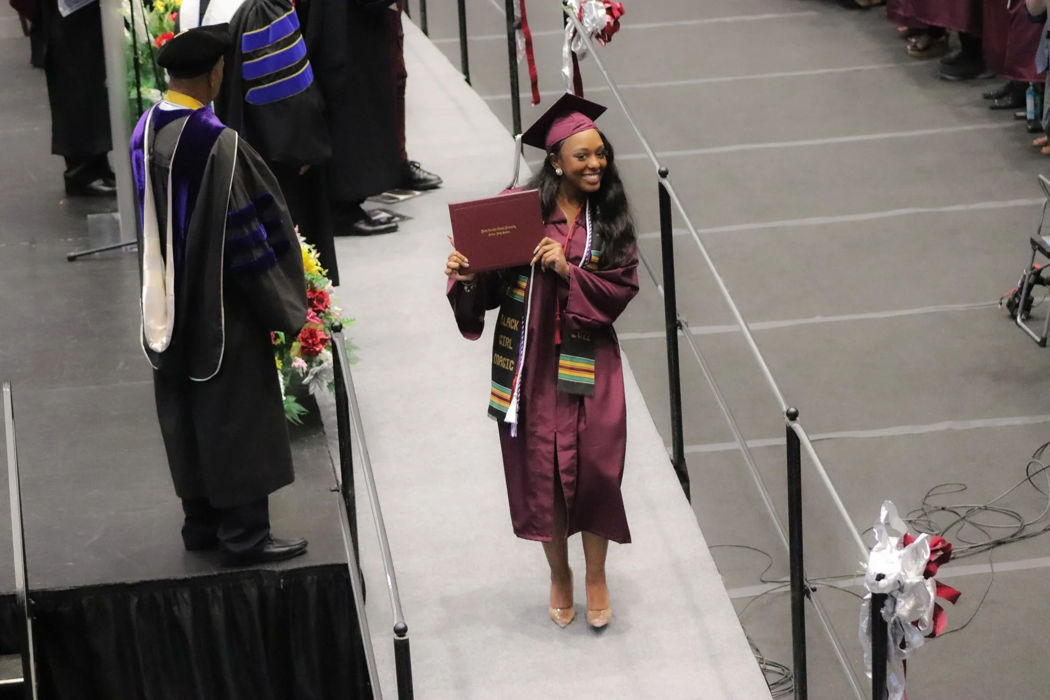 A young woman in a maroon graduation gown and cap smiling and holding her diploma while walking across the stage at her graduation ceremony. She is wearing a colorful stole and high heels.