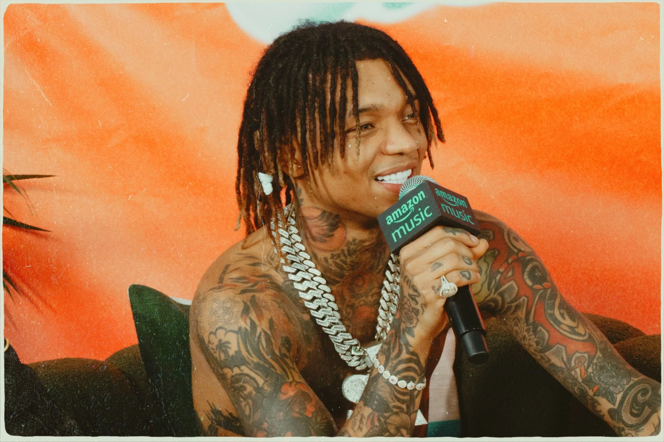A smiling woman with tattoos and dreadlocks holding a microphone labeled 'Amazon Music' during an interview or podcast.