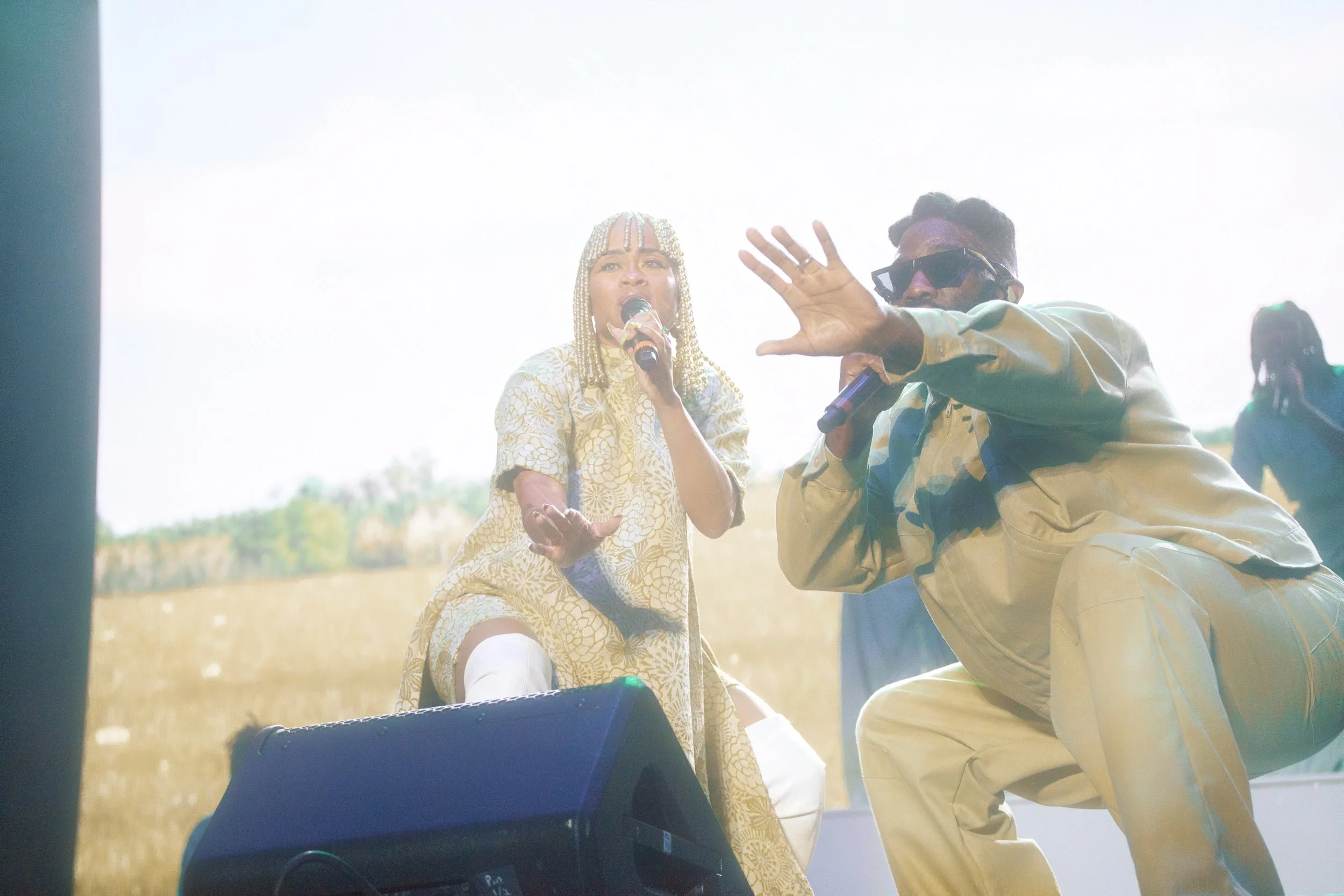 Two performers on stage, one woman dressed in gold with braided hair holding a microphone, and one man in beige outfit with sunglasses singing or speaking into a microphone.