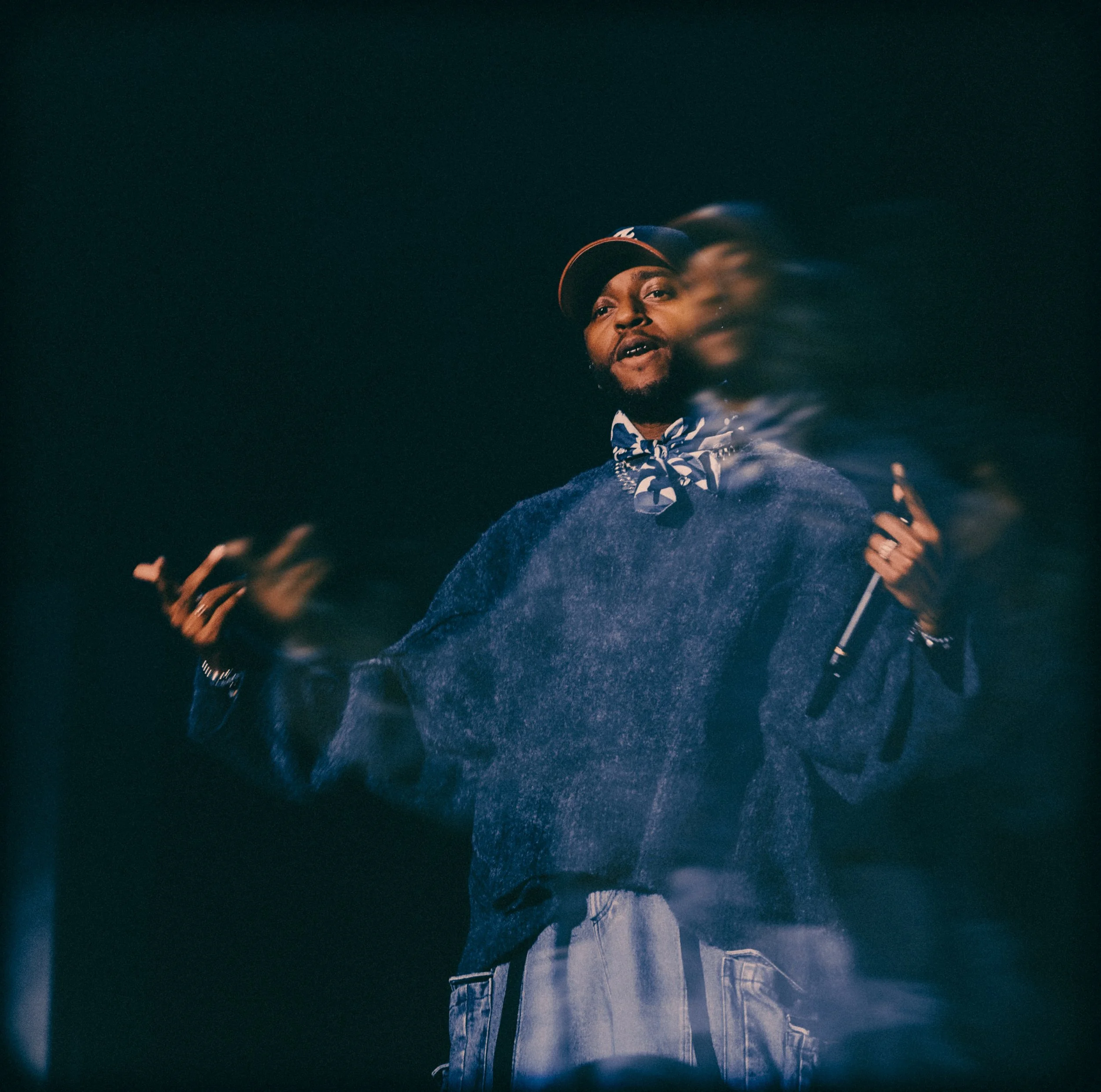 A man speaking on stage, wearing a sweater, bandana, baseball cap, and holding a microphone, with a blurred motion effect.