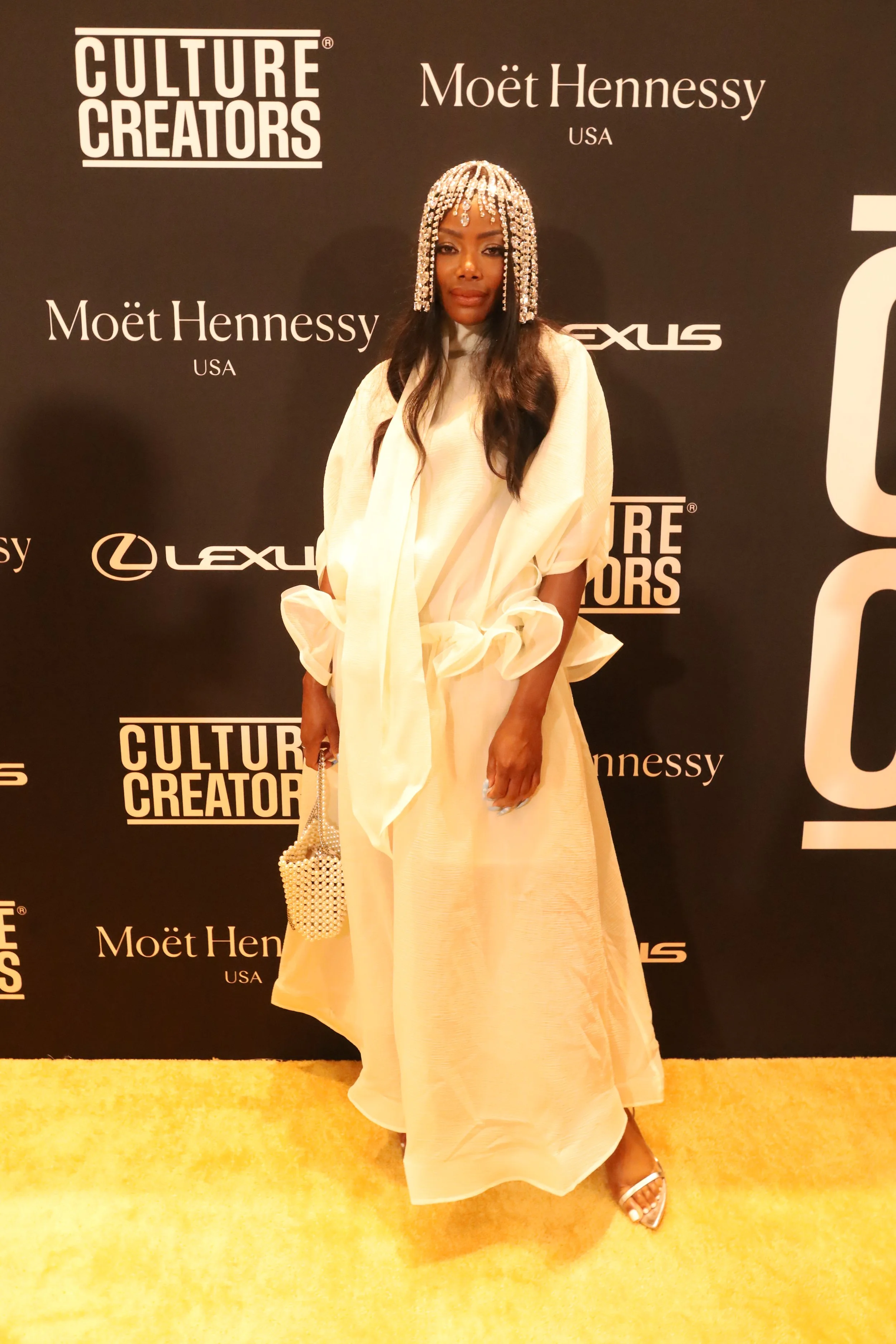 A woman in an elegant cream-colored dress with ruffled sleeves, standing on a yellow carpet at a formal event, holding a pearl-embellished handbag, and wearing a jewel-encrusted headpiece with long wavy dark hair.