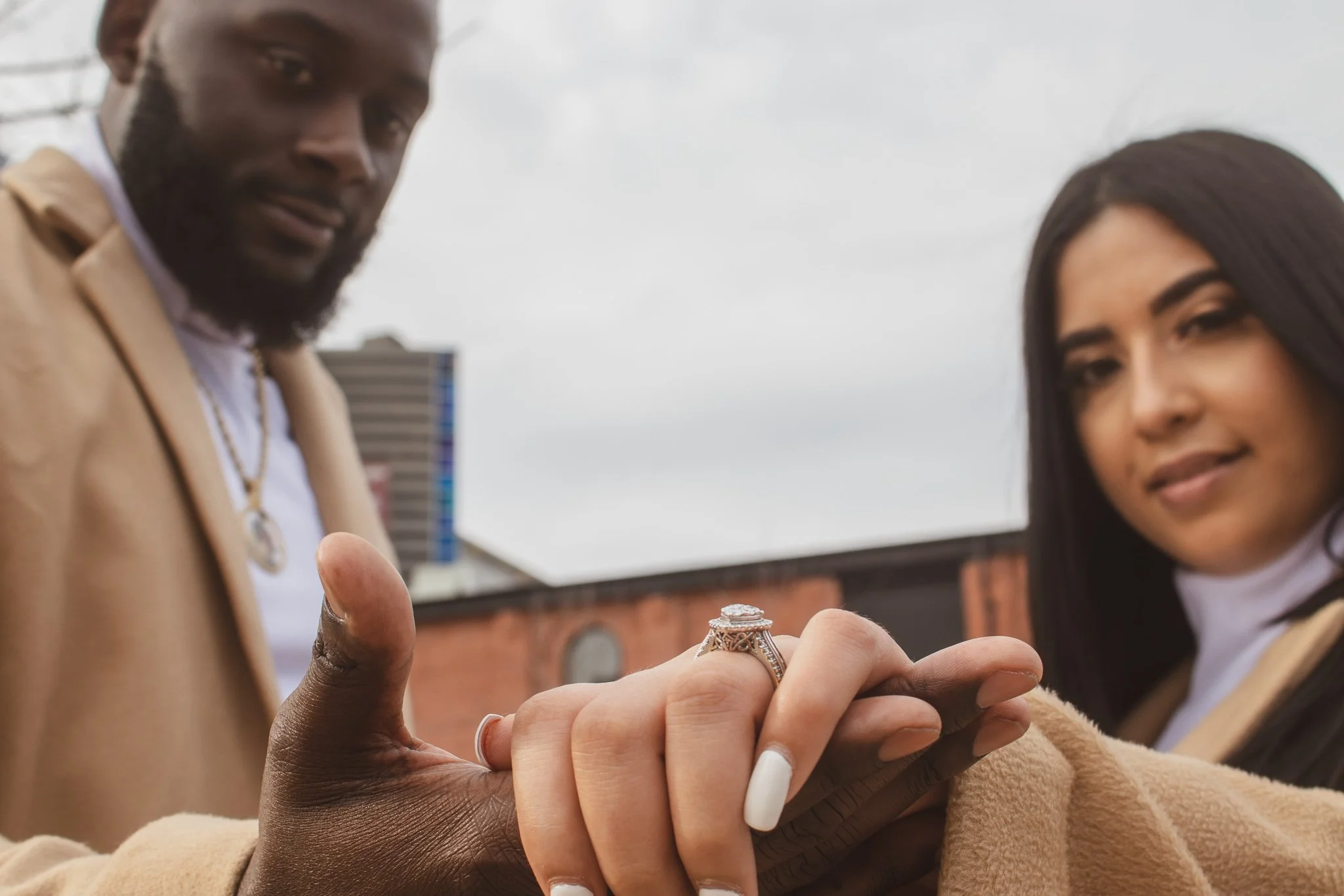 A woman and a man are holding hands, showing off a large engagement ring. The woman has long dark hair and is smiling, while the man has facial hair and is gazing at the camera. They are outdoors in an urban setting.
