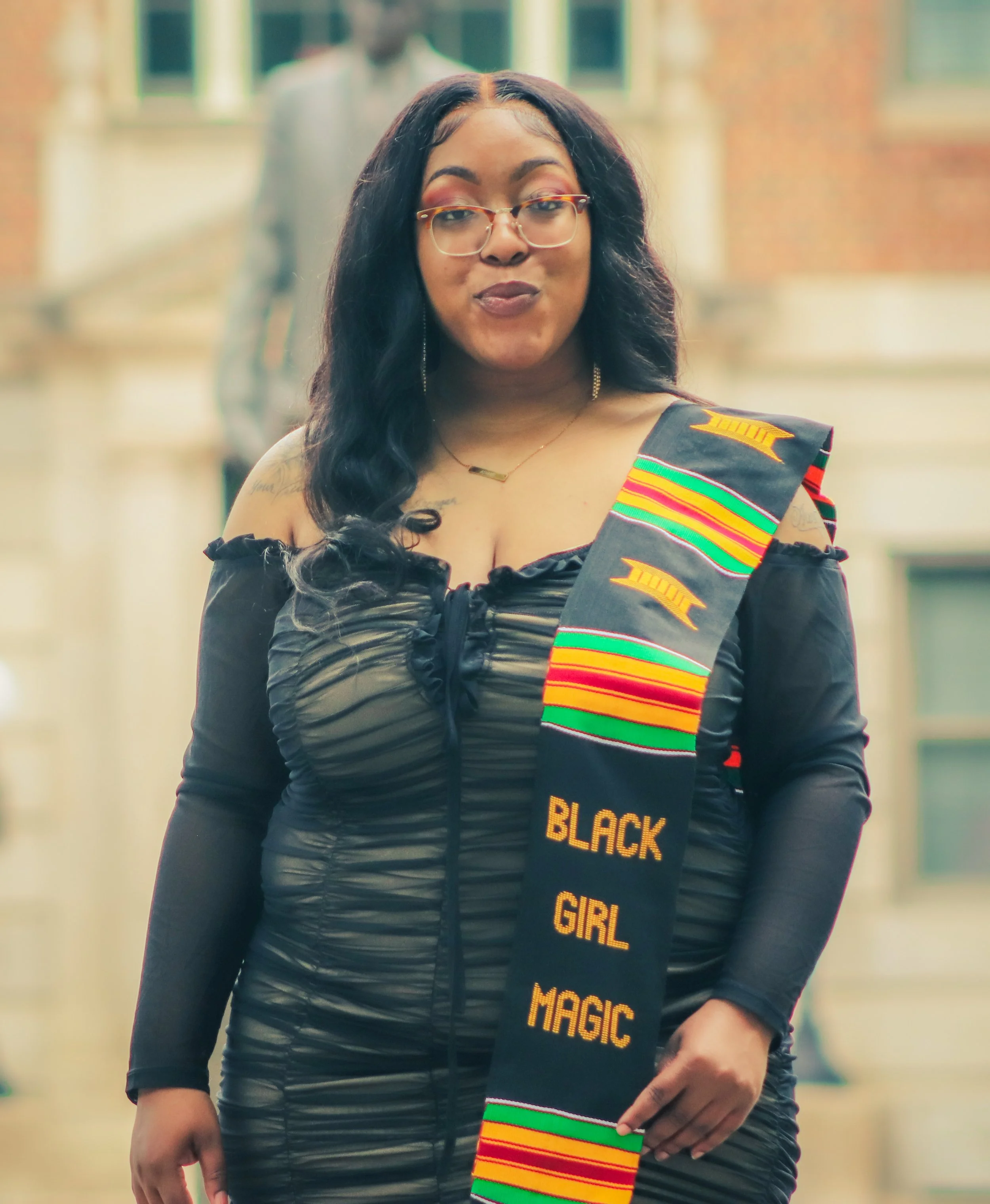 A young African American woman with long dark hair and glasses, wearing an off-shoulder black dress and a Kente cloth sash with the words "Black Girl Magic" embroidered on it.