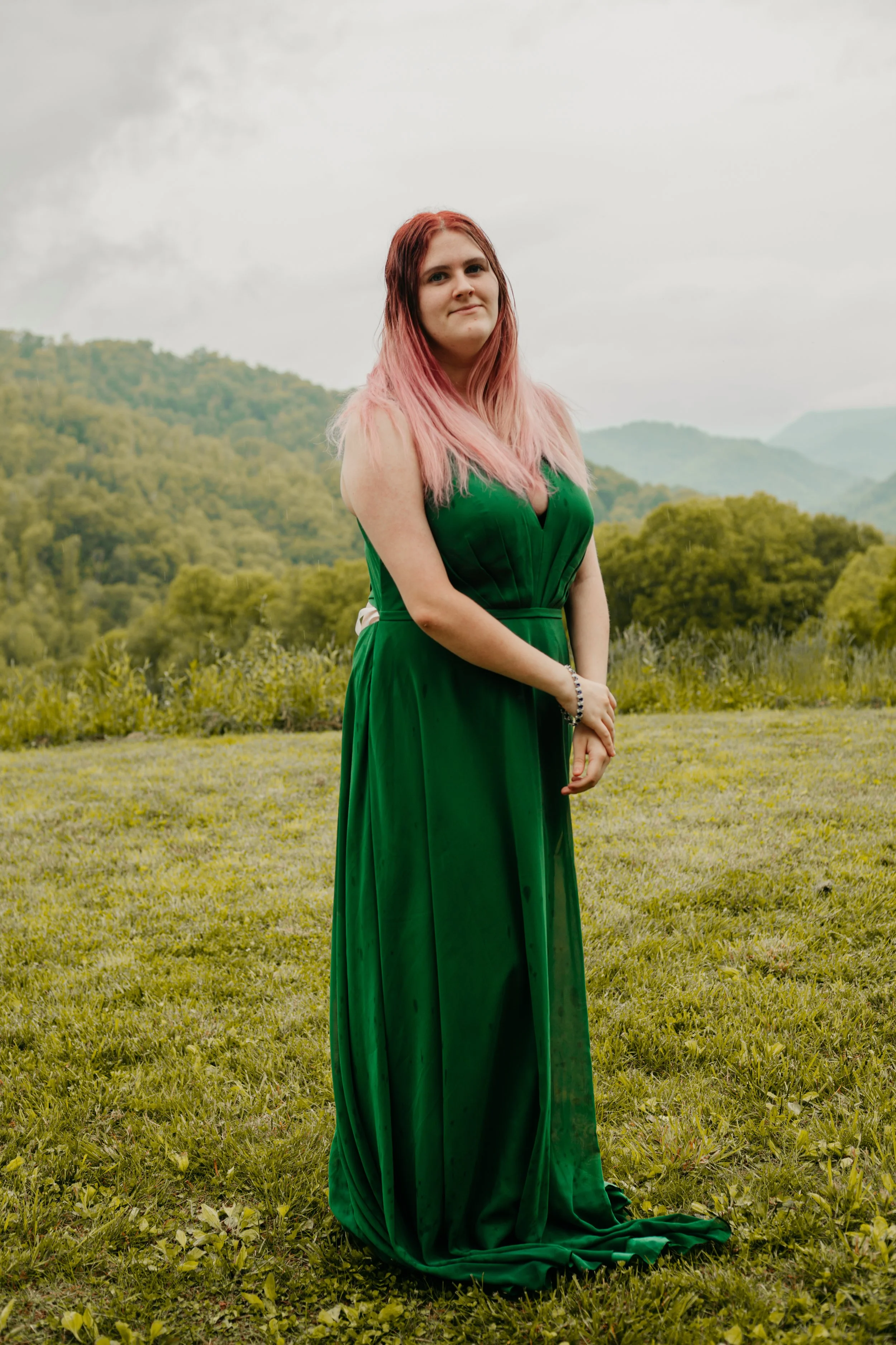 A woman with pink hair in a green dress standing outdoors on grass with hills and trees in the background.