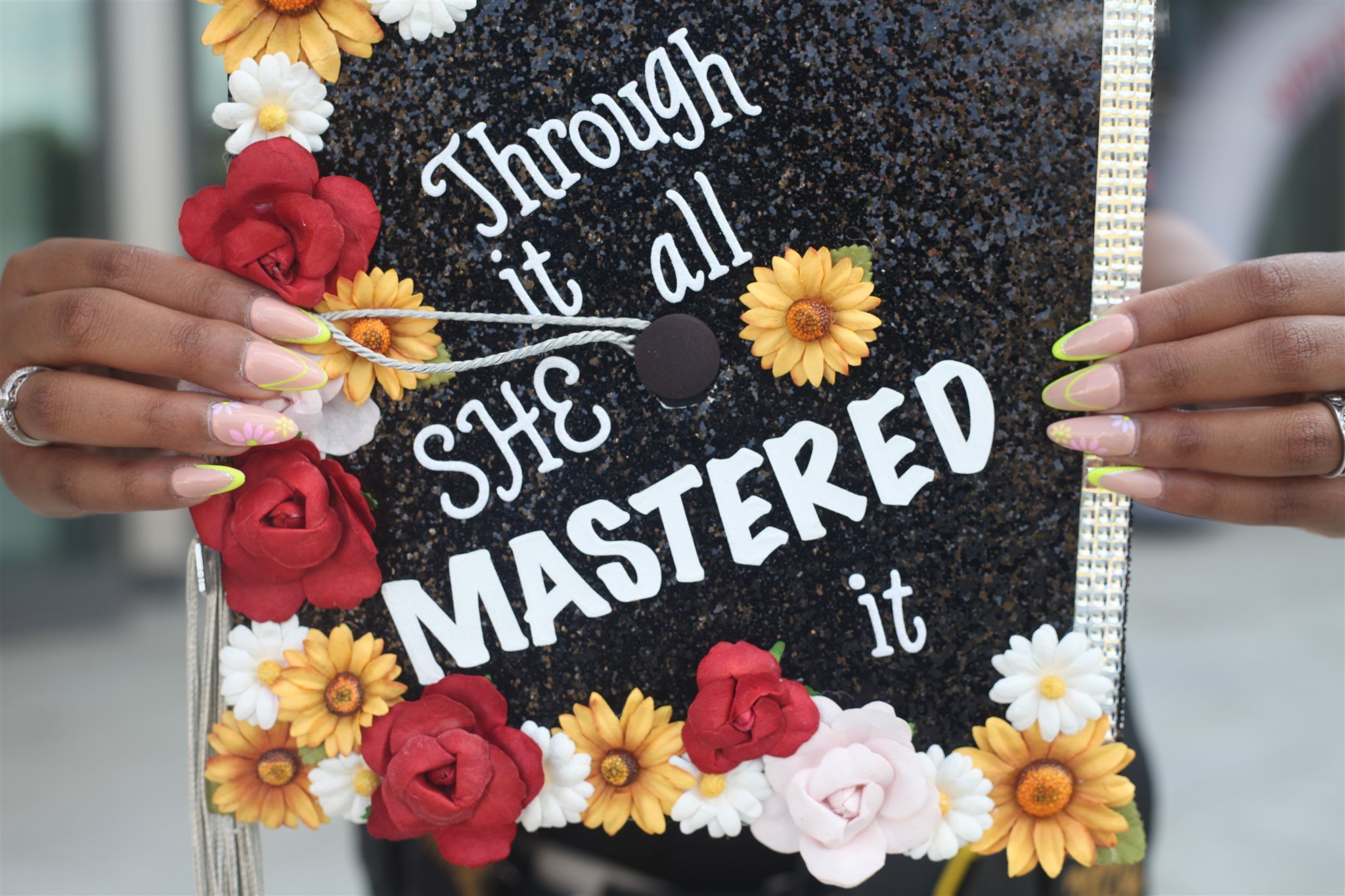 Hands holding a decorated graduation cap with the message 'Jrough it all says MASTERED,' surrounded by artificial flowers.