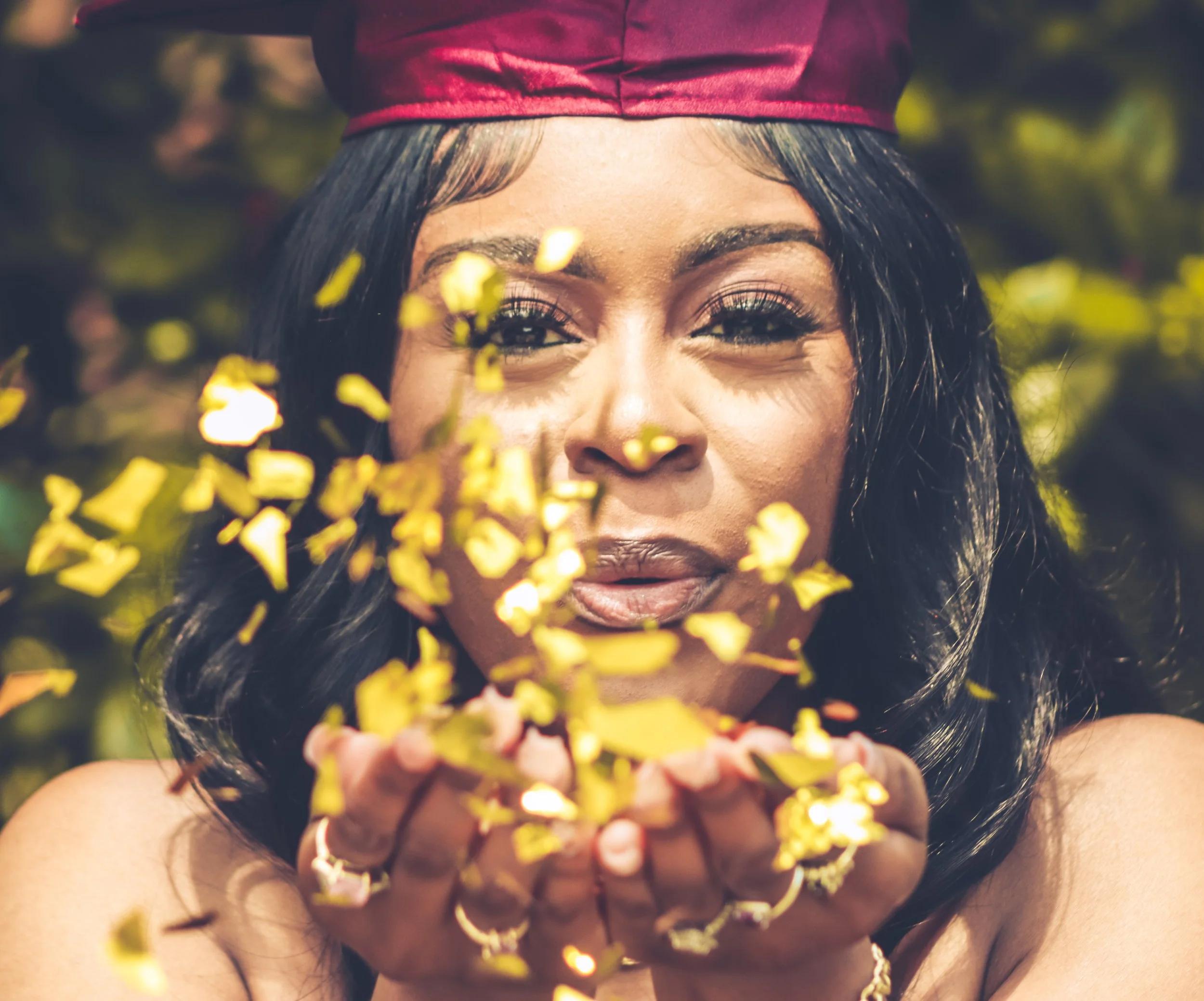 A woman wearing a graduation cap blows yellow confetti from her hands.