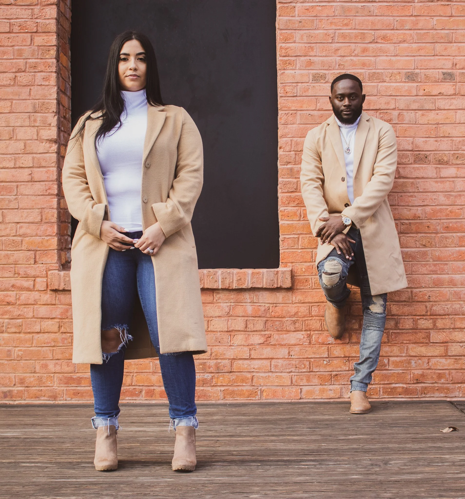 Two young adults, a woman and a man, are standing outdoors against a brick wall. The woman, on the left, is wearing a beige coat, white turtleneck, ripped jeans, and tan boots, with long dark hair. The man, on the right, is wearing a similar beige co