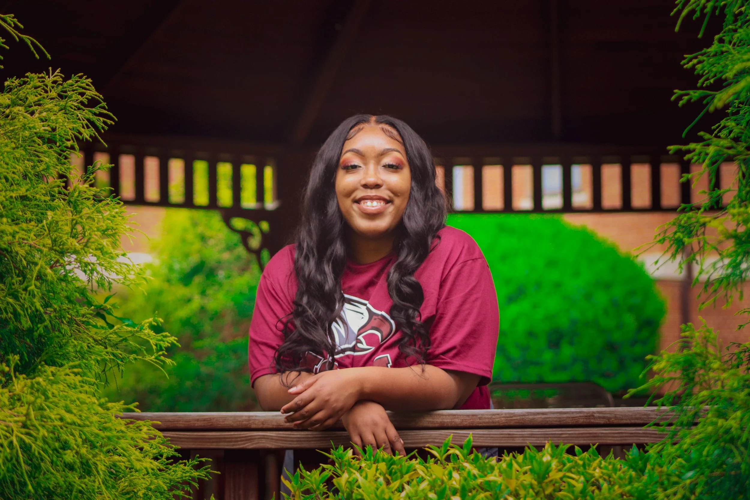 A young woman with long, wavy black hair smiling and leaning on a wooden railing surrounded by greenery, with a blurred background of trees and a brick structure.